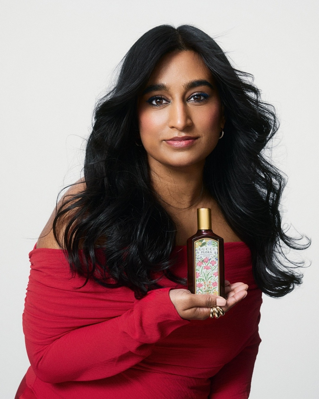 Eine Frau mit dunklen, gewellten Haaren und rotem Oberteil hält eine Flasche Gucci Flora mit einem goldenen Deckel und einem Etikett mit Blumenmuster.