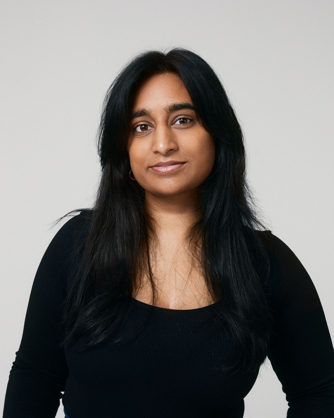 Porträt einer Frau mit dunklen, langen Haaren und einem schwarzen Oberteil vor einem neutralen Hintergrund.
