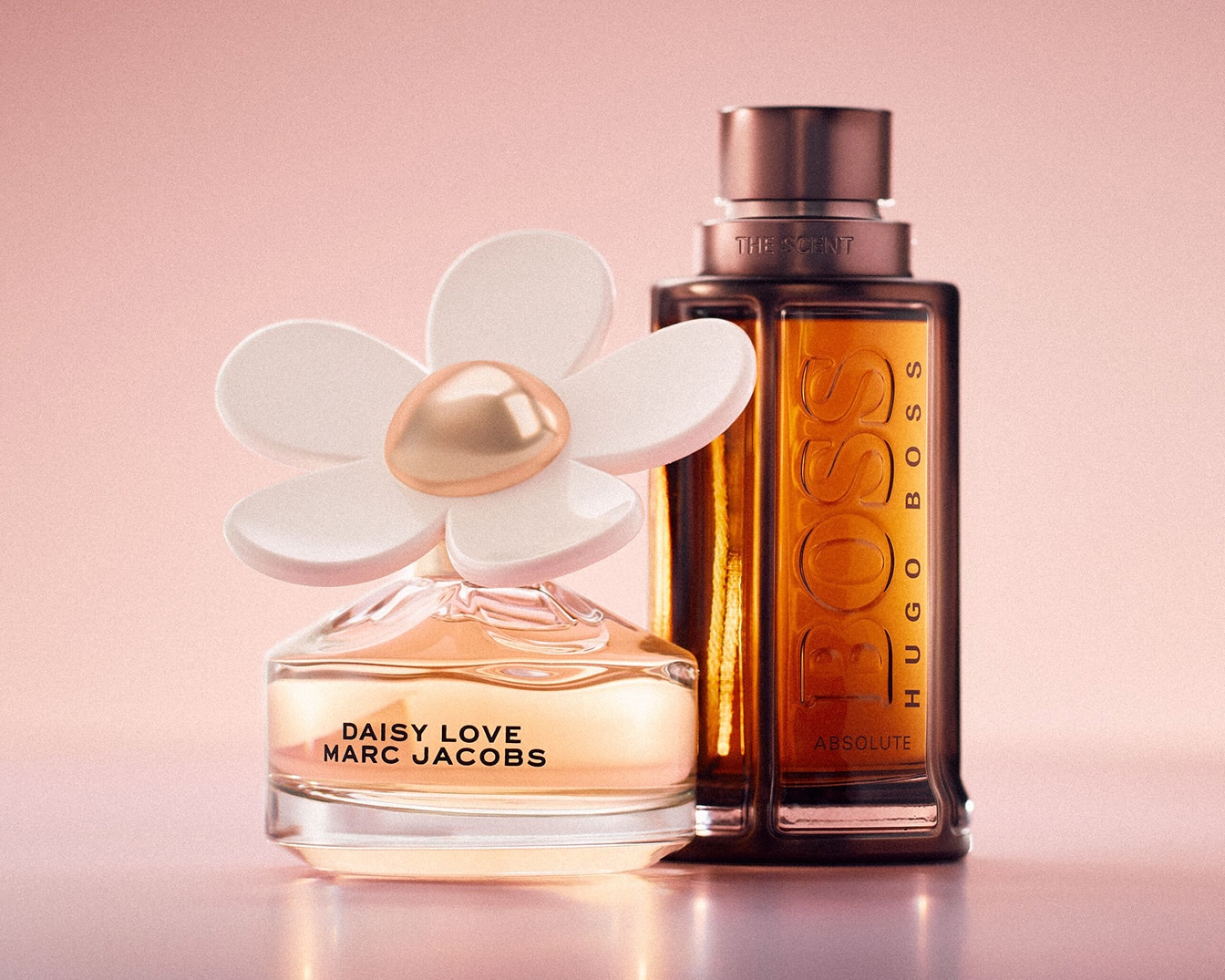 Na tle różu, Daisy Love Marc Jacobs z kwiatowym korkiem i Hugo Boss Absolute, oba flakony wypełnione płynem o ciepłym, bursztynowym odcieniu.