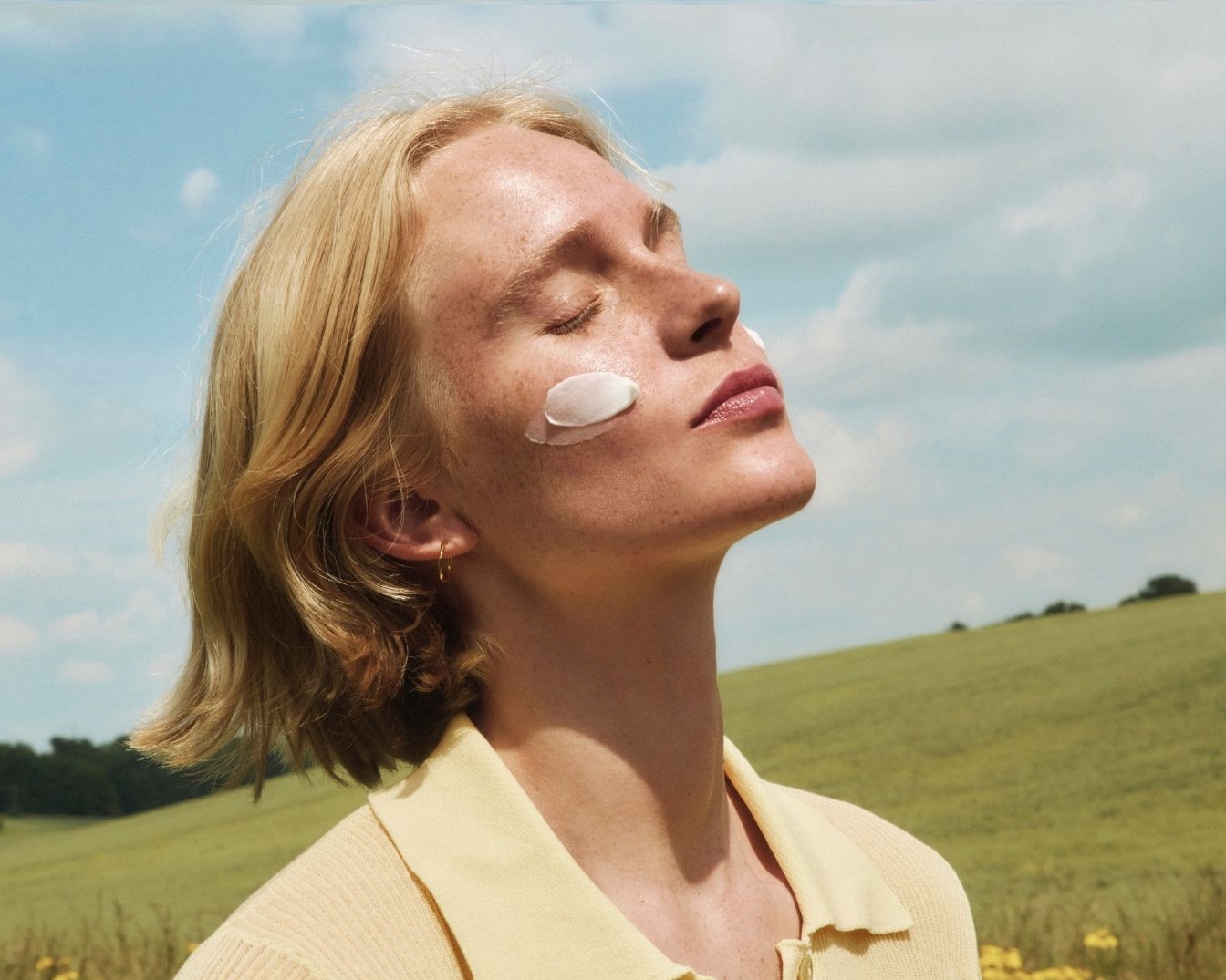 Blond kobieta z piegami, z zamkniętymi oczami, odchyla głowę do tyłu, na policzku i czole ma nałożony krem.