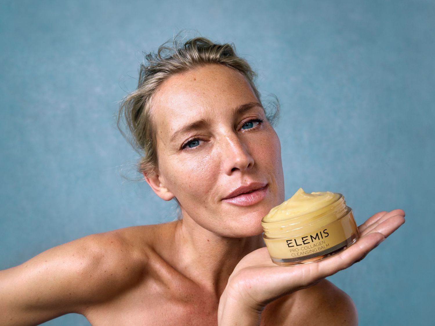 Žena drží v dlani sklenici s čisticím balzámem Elemis Pro-Collagen, který má žlutou barvu a krémovou texturu.