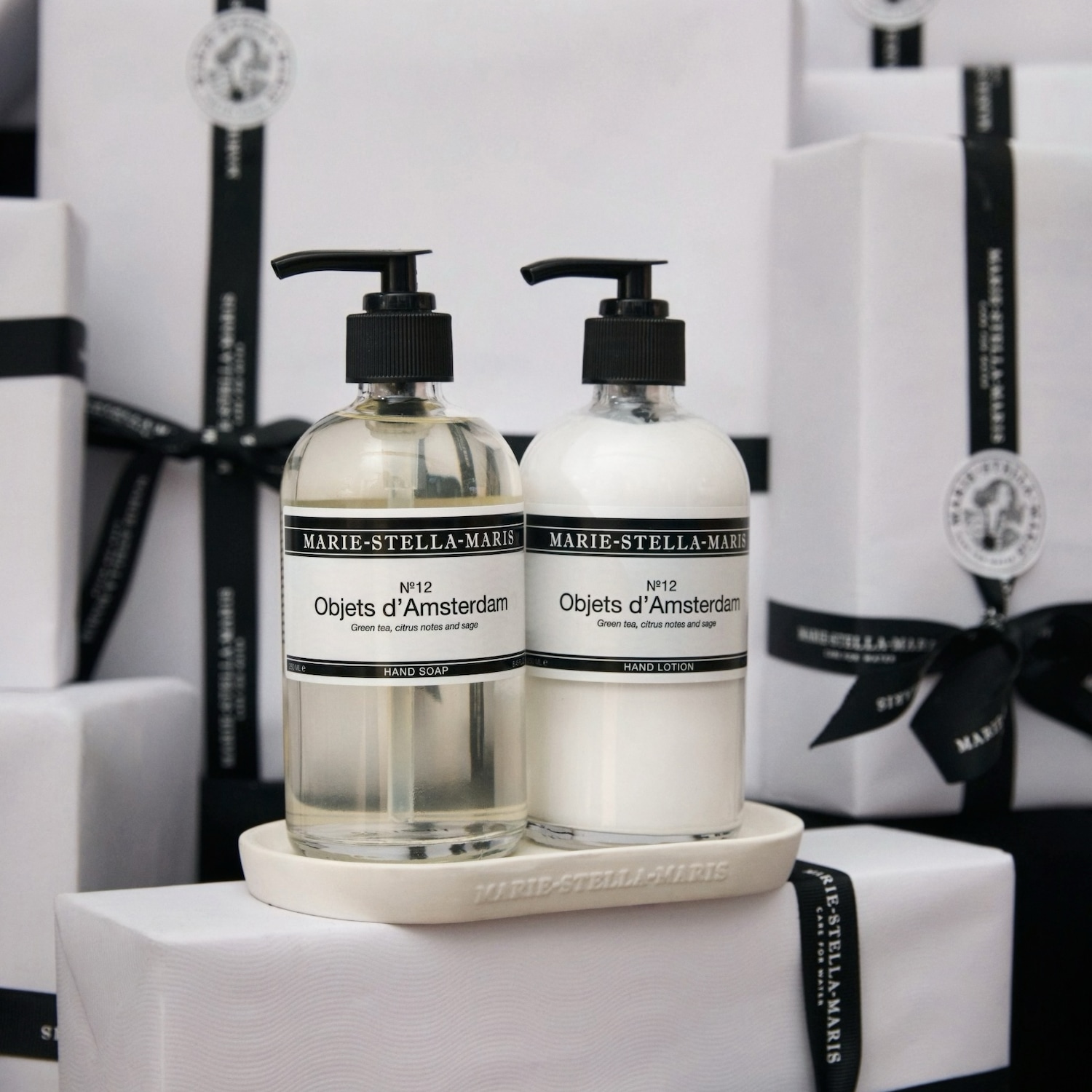 Twee flessen Marie-Stella-Maris handzeep en handlotion, 'No.12 Objets d'Amsterdam', met groene thee, citrus en salie, op een witte houder, omringd door witte geschenkdozen met zwarte linten.