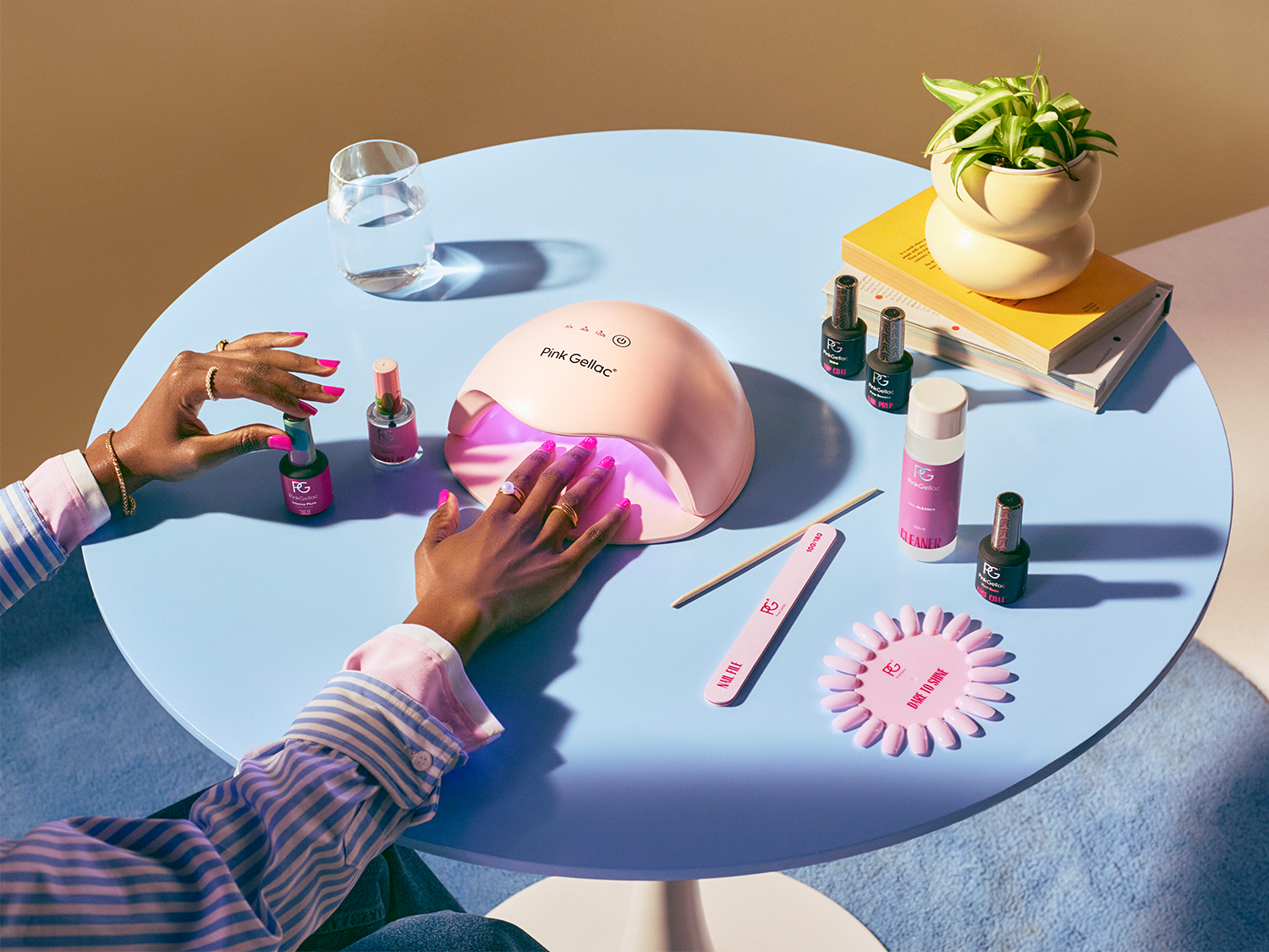 Une table bleu clair présente un ensemble de manucure Pink Gellac, avec une lampe UV rose allumée, des vernis à ongles colorés, une lime à ongles et une main appliquant du vernis rose vif.