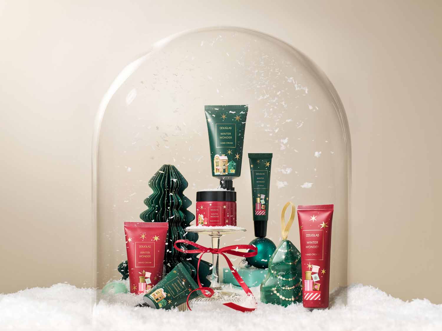 Kolekcja Douglas Winter Wonder w świątecznej scenerii pod szklaną kopułą, prezentująca zielone i czerwone opakowania kremów do rąk i balsamów do ust.