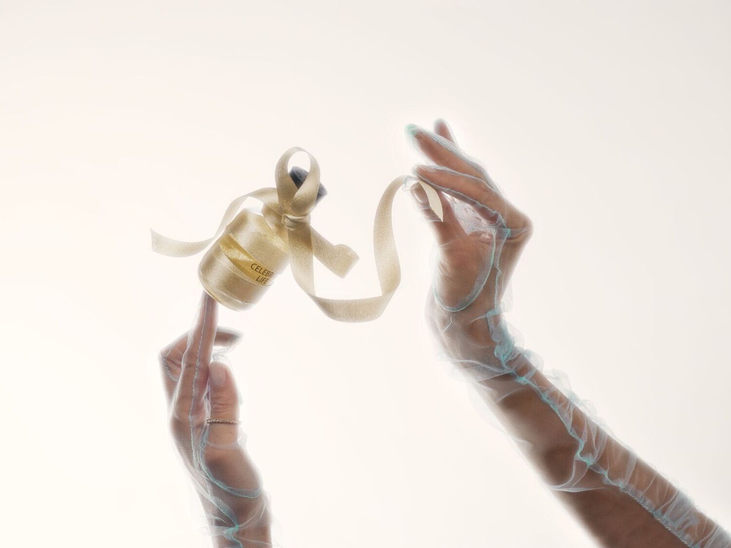 Švelniai apšviestas vaizdas, kuriame rankos su permatomomis pirštinėmis laiko mažą stiklinį buteliuką, aprištą auksiniu kaspinu, ant kurio užrašas „Švęsk gyvenimą“.