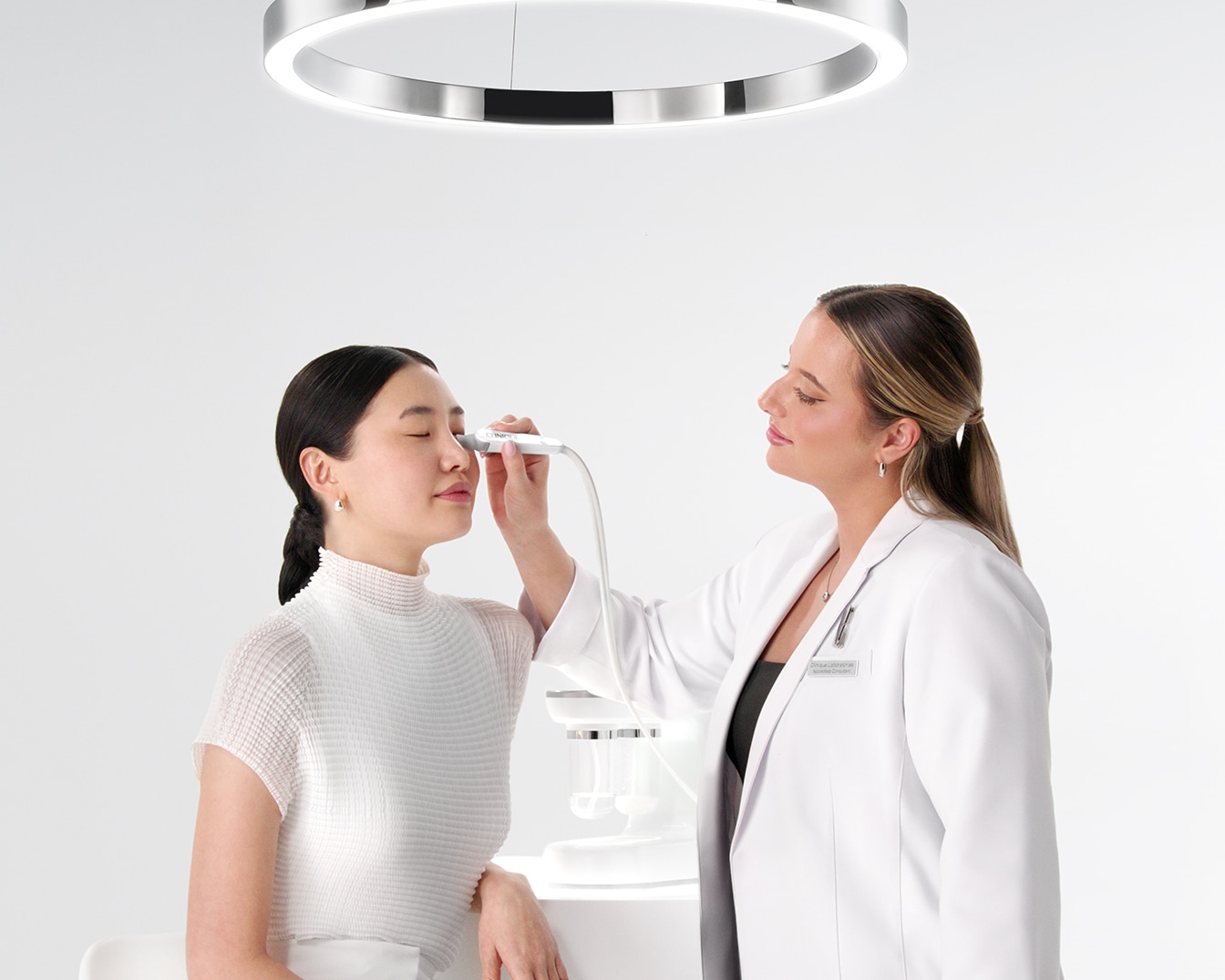 Una donna riceve un trattamento viso da un'estetista con un dispositivo bianco, sotto una luce circolare brillante.