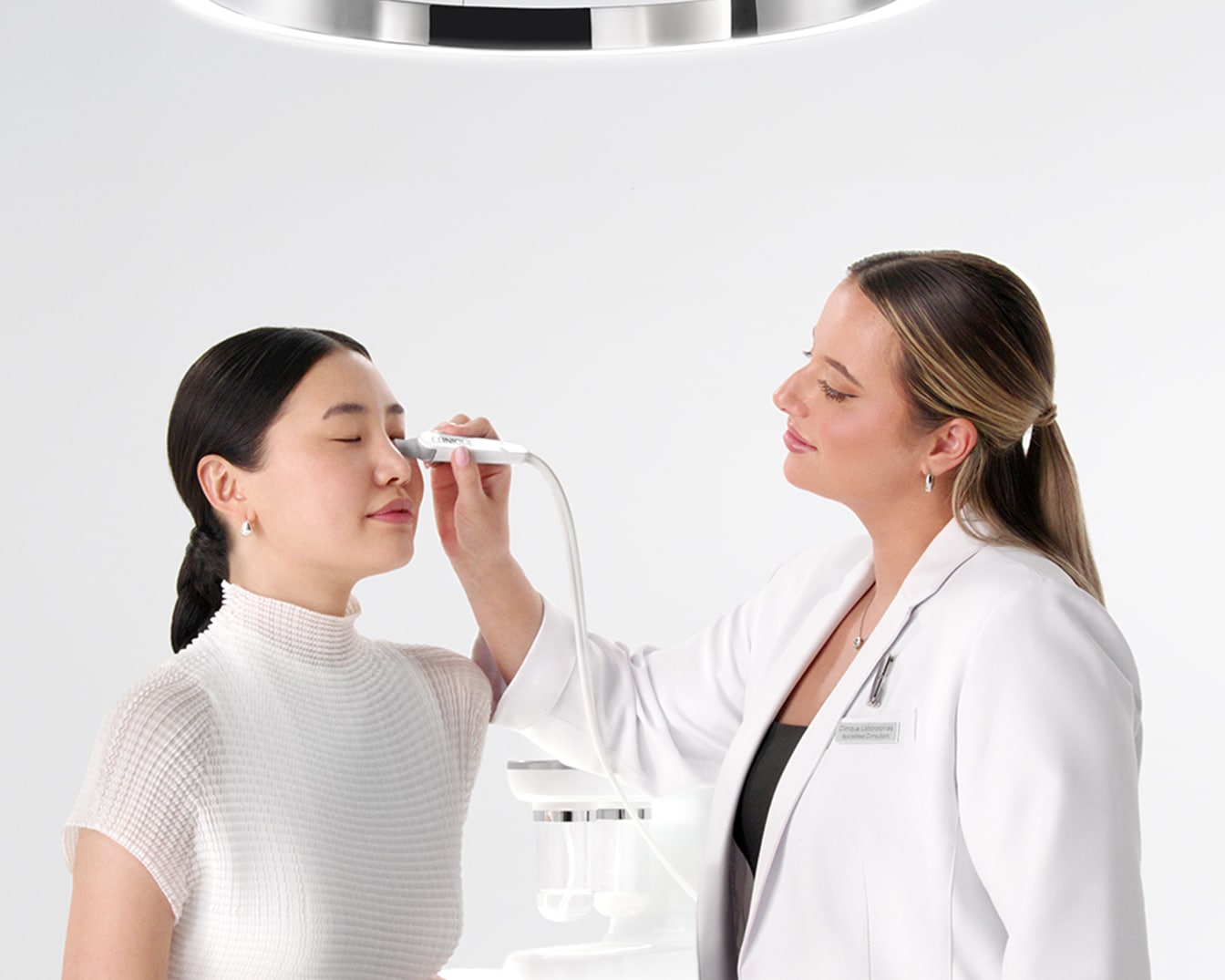 Un'estetista in camice bianco esegue un trattamento viso su una cliente con un dispositivo elettronico, in un ambiente luminoso e professionale.