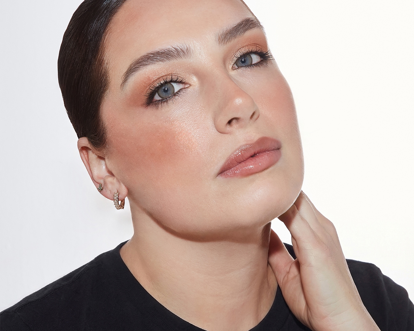 Primo piano di una donna con trucco luminoso sugli occhi, fard pesca e labbra lucide, che mette in risalto la sua bellezza naturale.