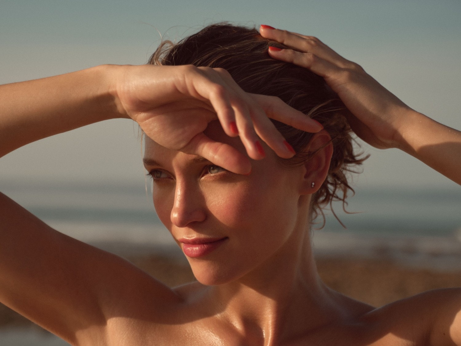 Eine Frau mit nassen, gewellten Haaren und roten Nägeln hält ihre Hände über ihren Kopf, um sich vor der Sonne zu schützen, während sie am Strand steht.
