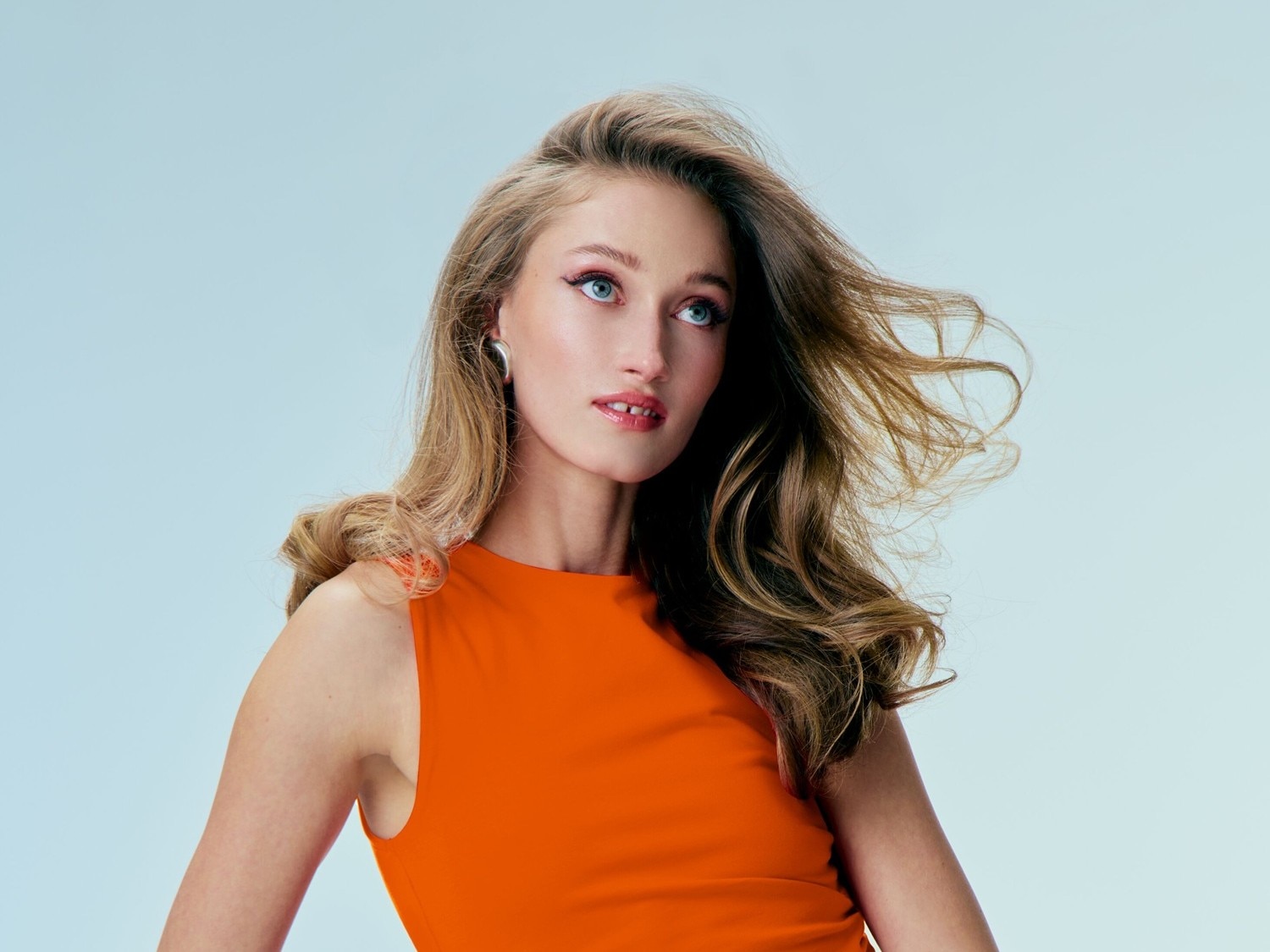 Eine Frau mit gewelltem, blondem Haar und einem orangefarbenen Kleid blickt mit strahlenden blauen Augen nach oben.