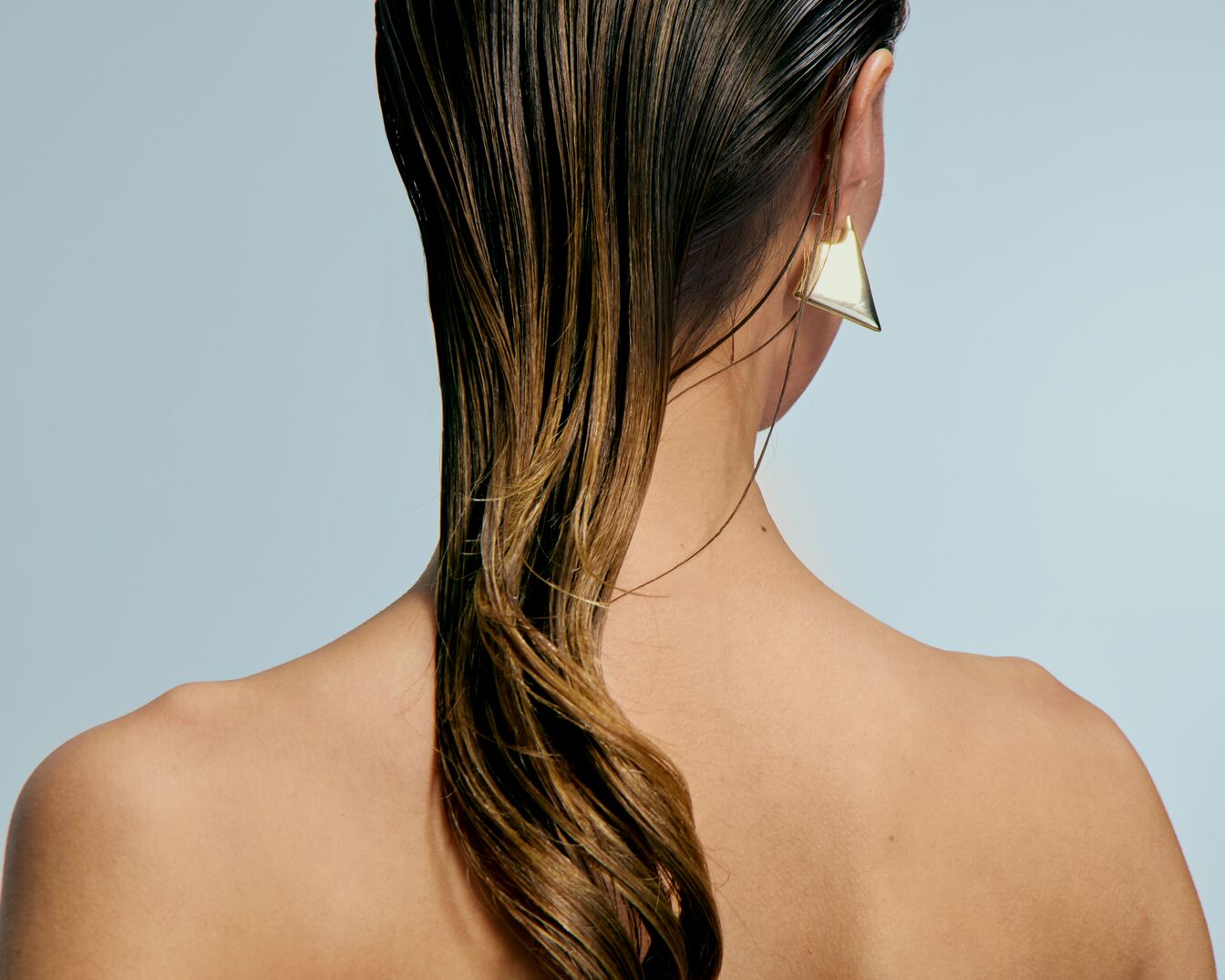 Nahaufnahme einer Frau mit nassen, braunen Haaren, die über ihre Schulter fallen, und einem goldenen, dreieckigen Ohrring.