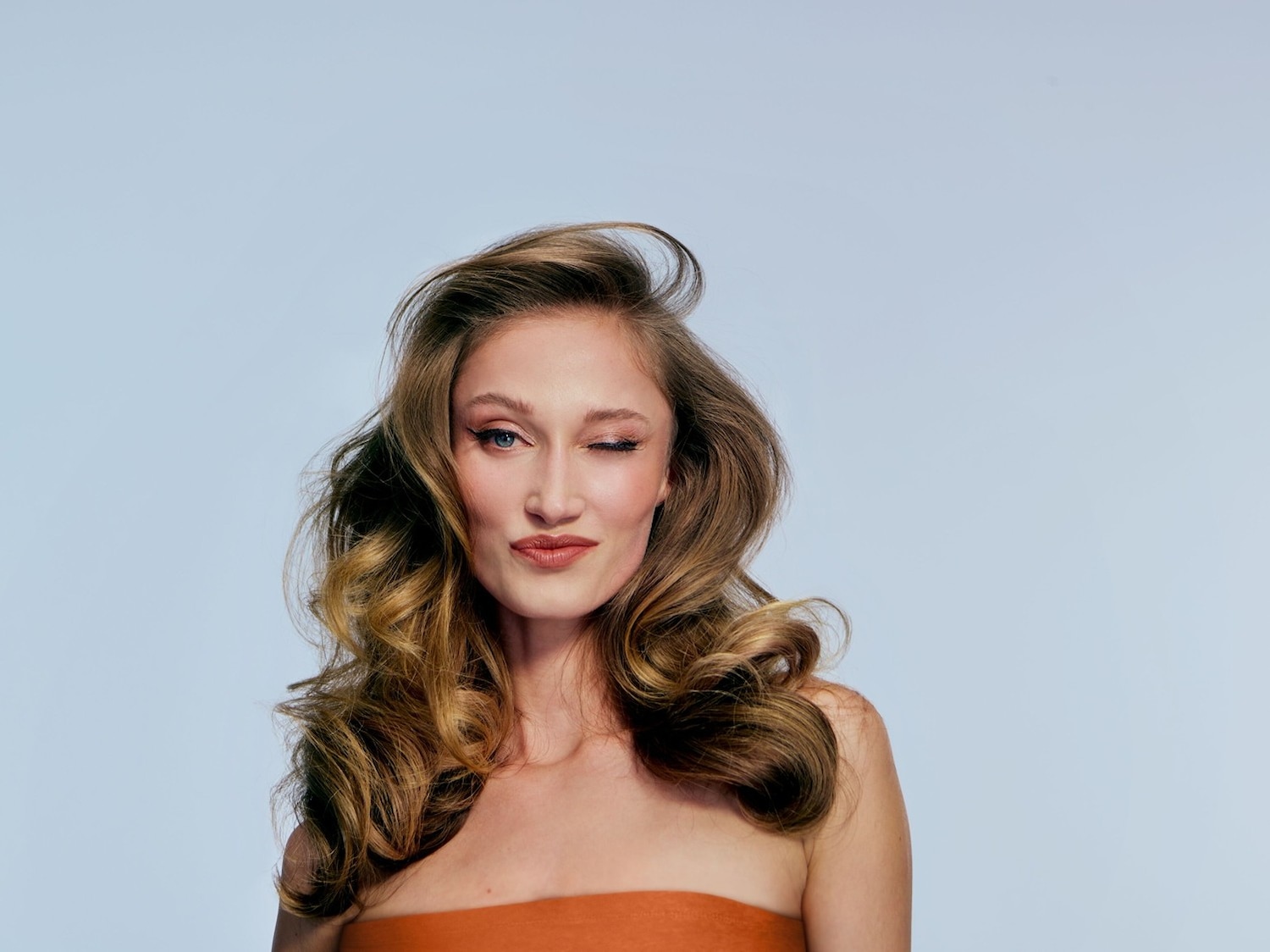 Porträt einer Frau mit gewelltem, braunem Haar, die ein orangefarbenes trägerloses Oberteil trägt und mit einem Auge zwinkert.