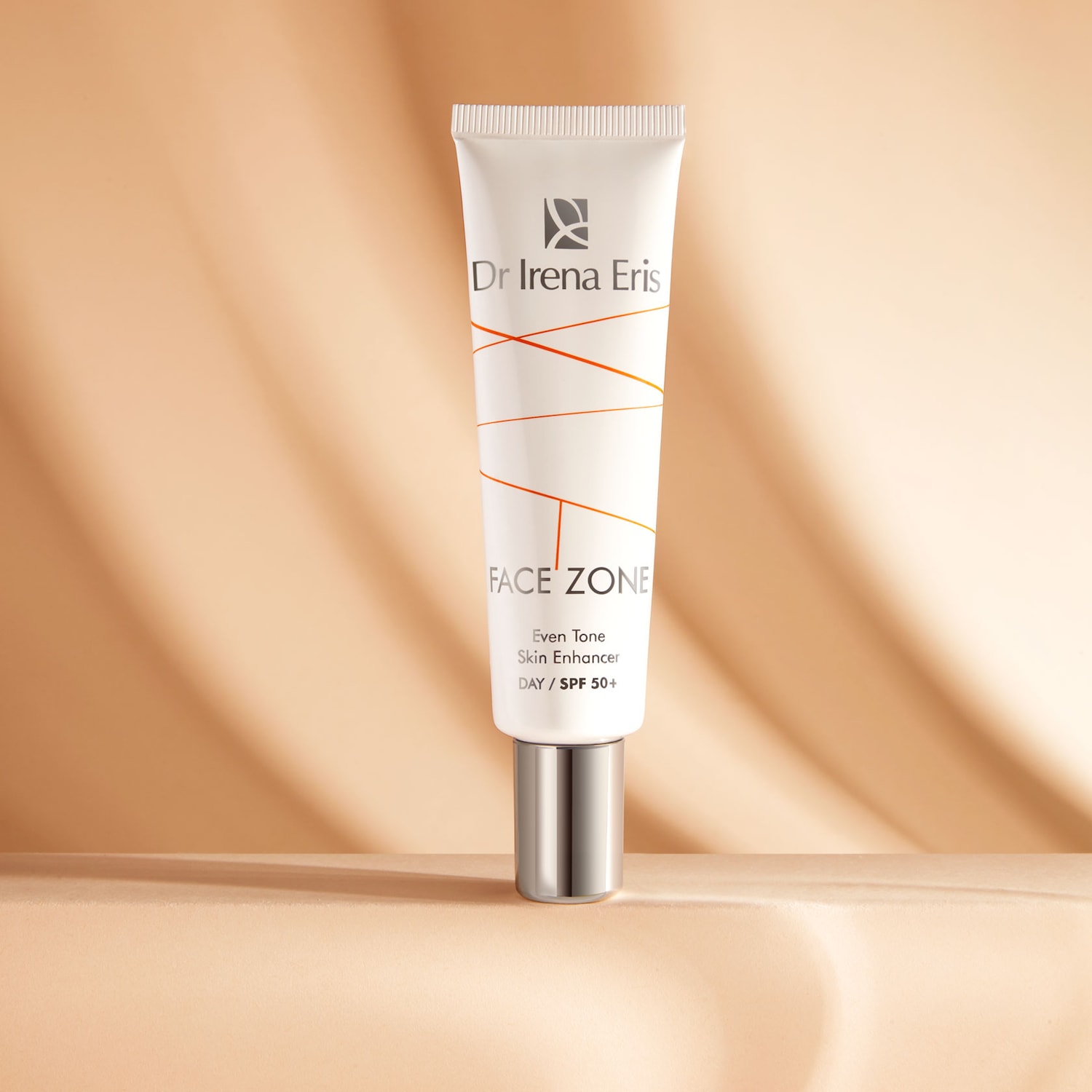 Biała tuba kremu Dr Irena Eris Face Zone Even Tone Skin Enhancer DAY/SPF 50+ z pomarańczowymi liniami na beżowym tle.