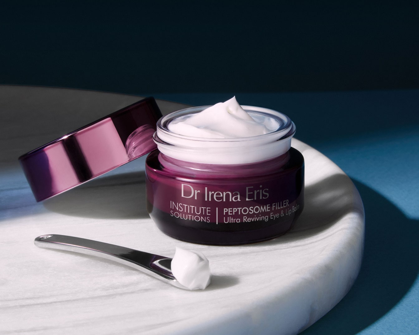 Dr Irena Eris Peptosome Filler Ultra Reviving Eye & Lip Balm w fioletowym słoiczku z białą zawartością i srebrną szpatułką na marmurowej powierzchni.