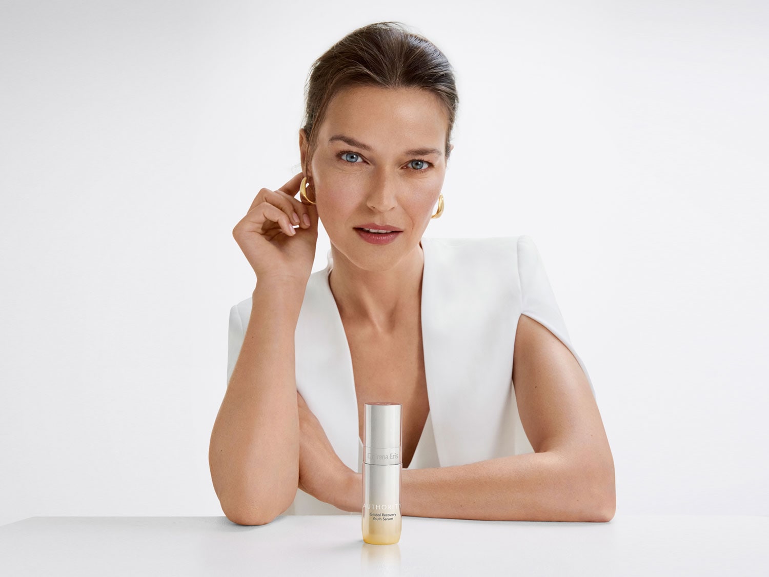 Na pierwszym planie kobieta z niebieskimi oczami i złotymi kolczykami, ubrana w białą marynarkę, obok niej stoi srebrna butelka serum do twarzy.