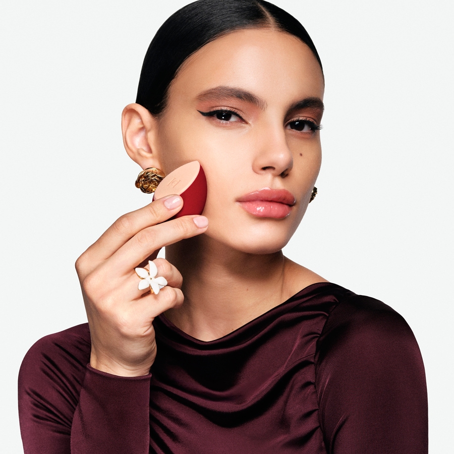 Eine Frau mit dunklen Haaren und einem weinroten Oberteil trägt mit einem roten Make-up-Schwamm Foundation auf ihr Gesicht auf.