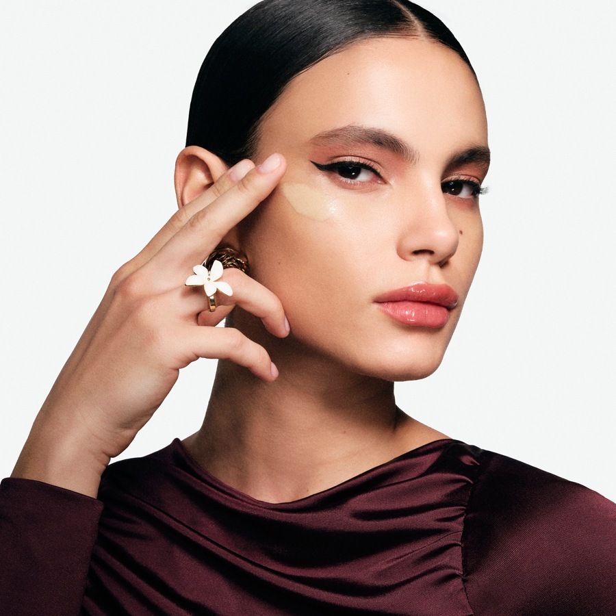 Nahaufnahme einer Frau, die mit einem Finger eine hellbeige Creme unter dem Auge aufträgt, mit einem goldenen Ring mit einer weißen Blume am Finger.