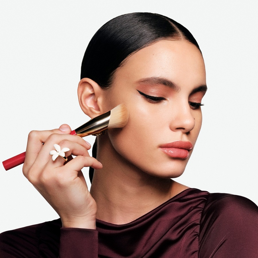 Eine Frau mit glatten, dunklen Haaren und einem weinroten Oberteil trägt mit einem Pinsel Make-up auf ihr Gesicht auf.