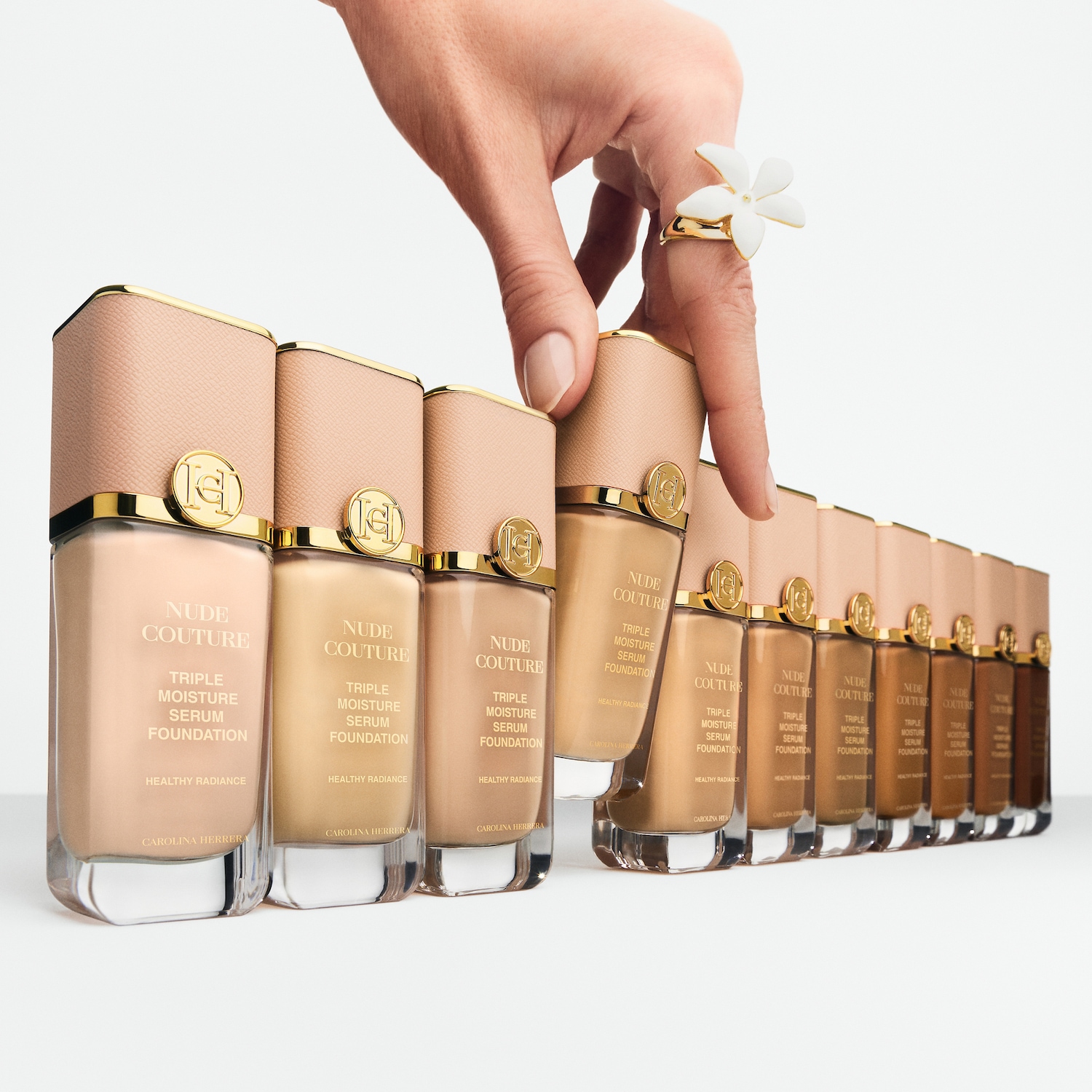 Eine Hand mit einem Blumenring greift nach Carolina Herrera Nude Couture Triple Moisture Serum Foundation, erhältlich in verschiedenen Farbtönen für einen gesunden Glanz.