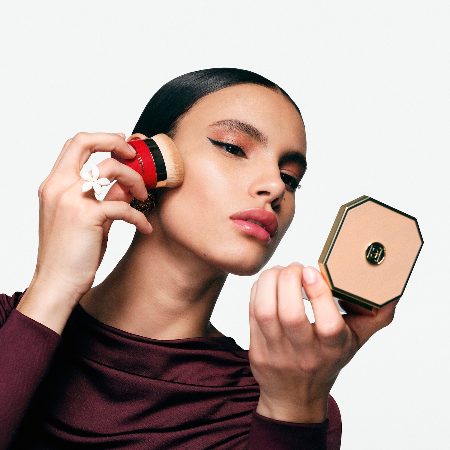 Eine Frau mit glatten, dunklen Haaren trägt ein weinrotes Oberteil und trägt mit einem Pinsel Make-up auf, während sie in einen goldenen Kompaktspiegel schaut.