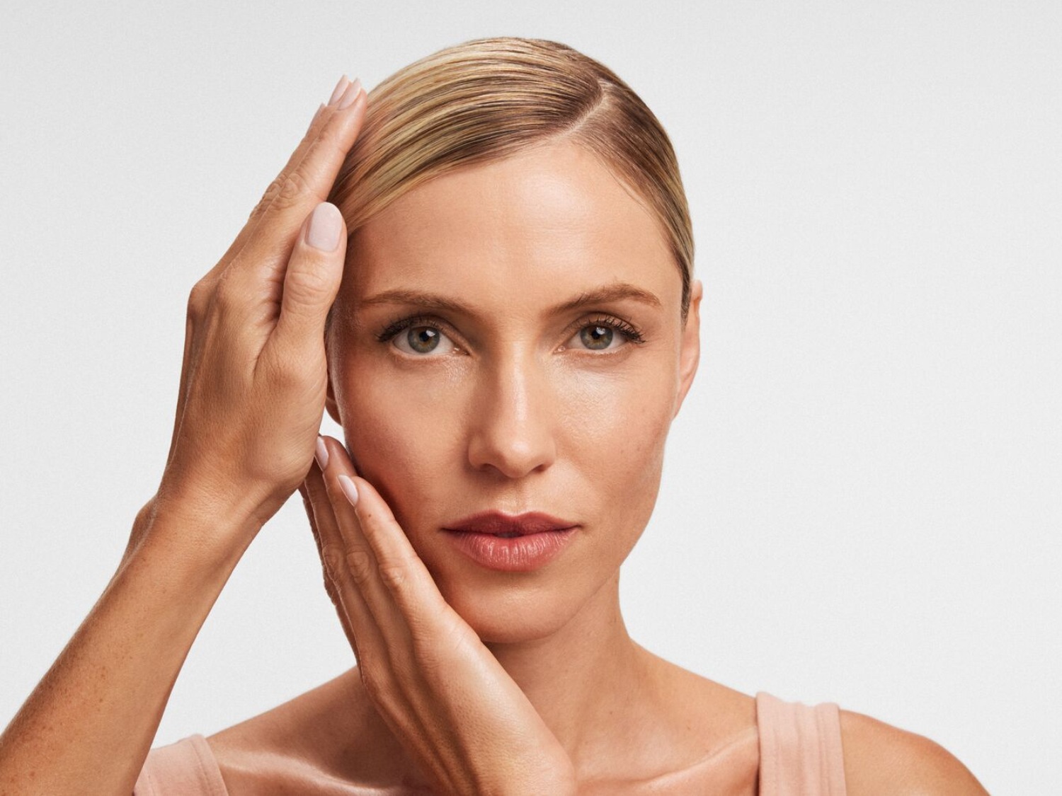 Egy szőke hajú nő, mindkét kezével az arcát érinti, miközben egy rózsaszín felsőt visel, és egyenesen a kamerába néz.