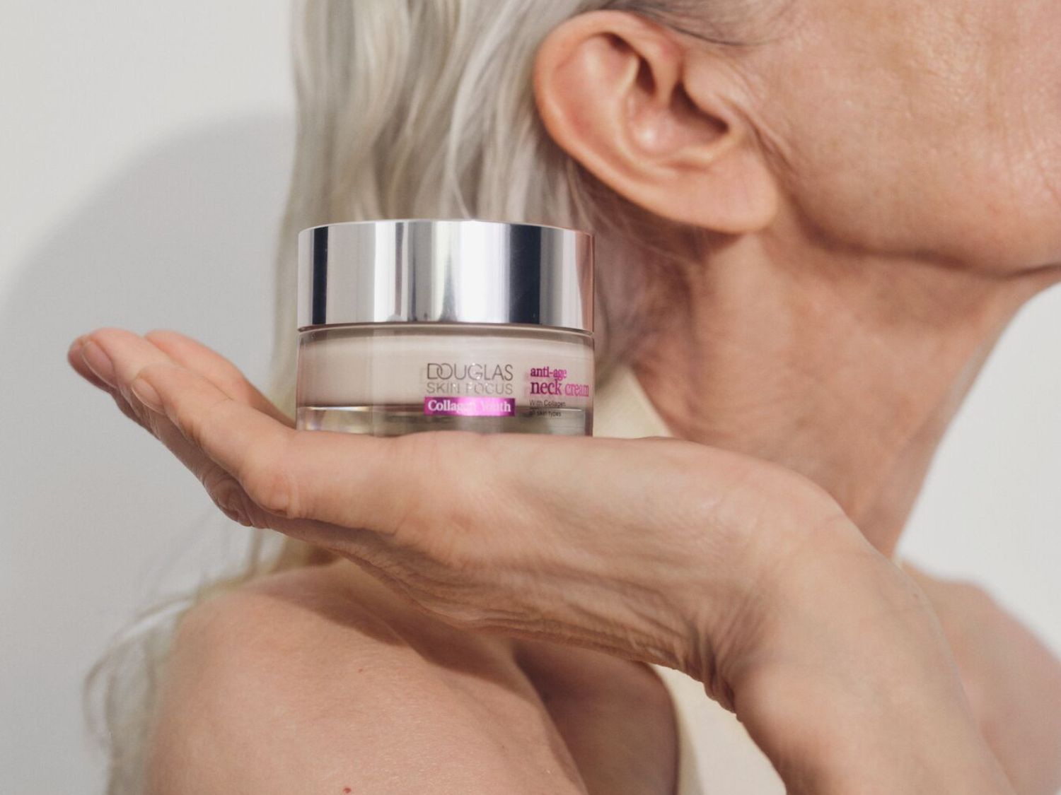 Egy nő tartja a kezében a Douglas Skin Focus Anti-Age nyakkrém tégelyét, amely kollagénnel gazdagított, hogy minden bőrtípus számára feszesebb nyakat biztosítson.