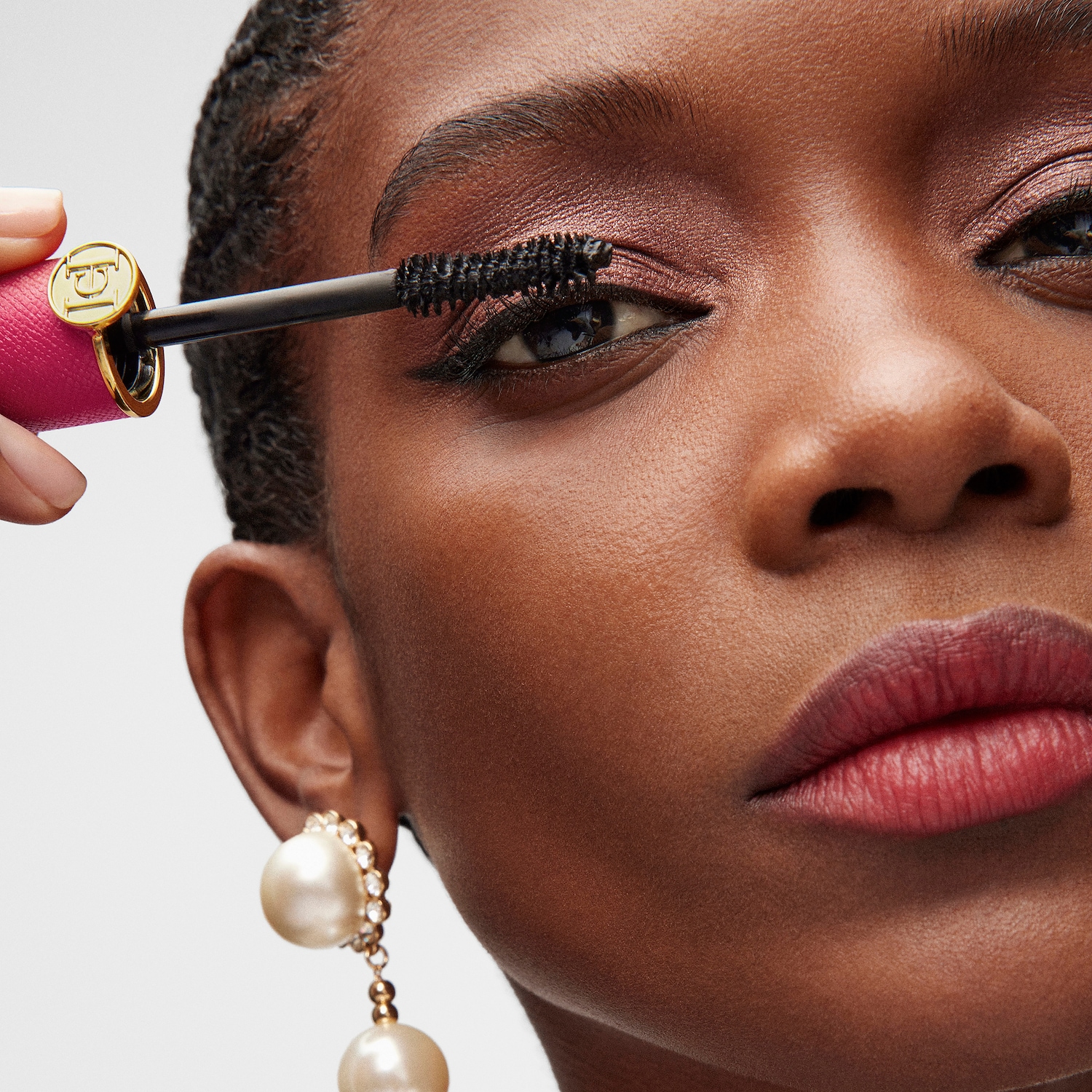 Nahaufnahme einer Frau, die sich mit einer pinkfarbenen Mascara die Wimpern tuscht, dazu passend rosafarbener Lidschatten und Ohrringe mit großen Perlen.