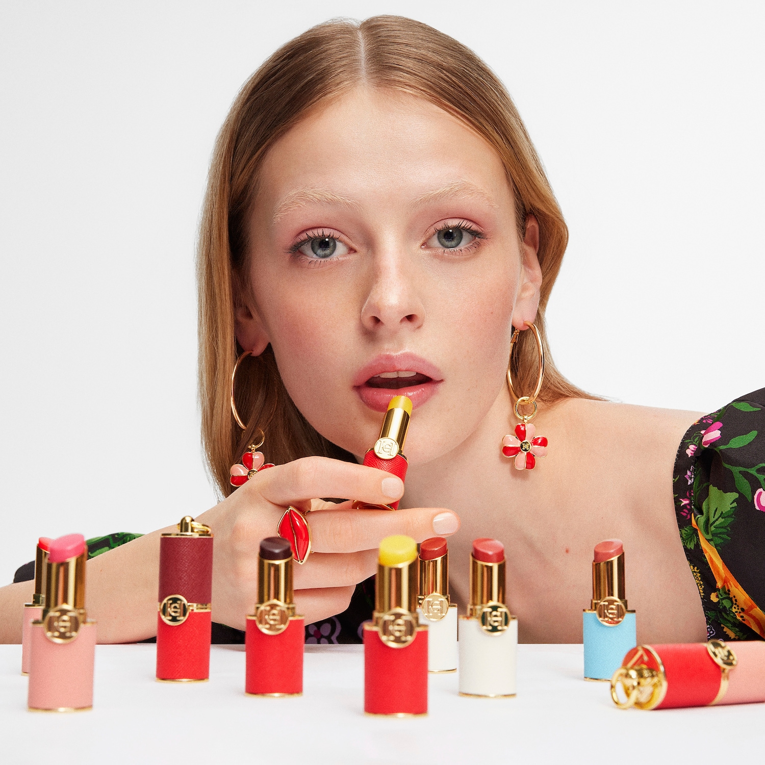 Eine Frau mit blonden Haaren trägt Ohrringe und einen Ring in Form einer roten Lippe, während sie einen gelben Lippenstift aufträgt, umgeben von einer Kollektion verschiedenfarbiger Lippenstifte.