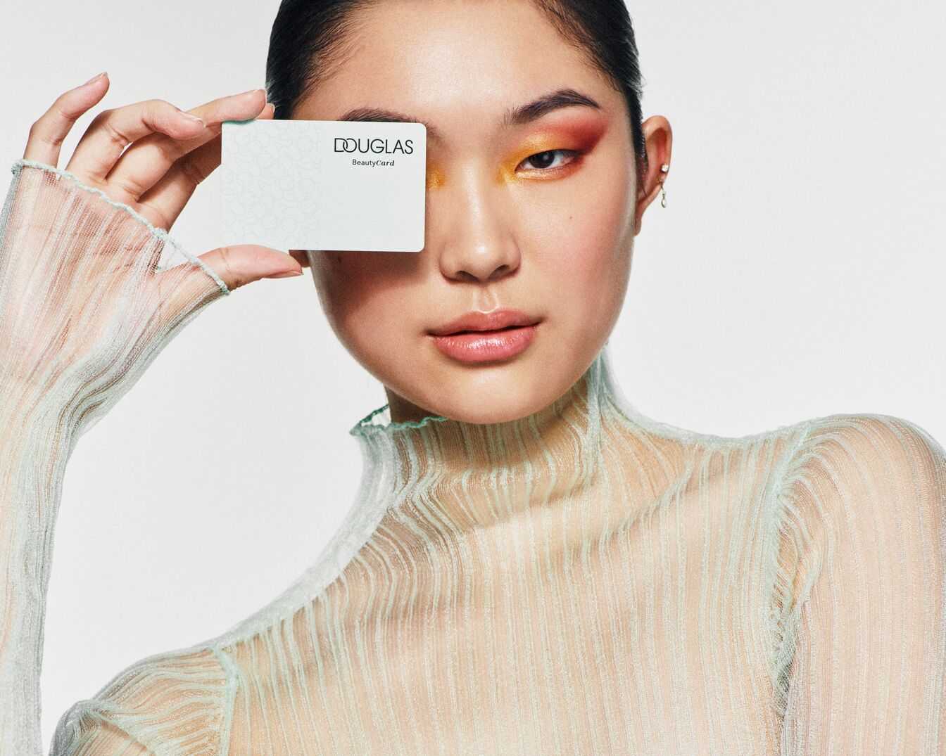 Portrét ženy s barevným očním make-upem, která drží kartu Douglas BeautyCard před okem.
