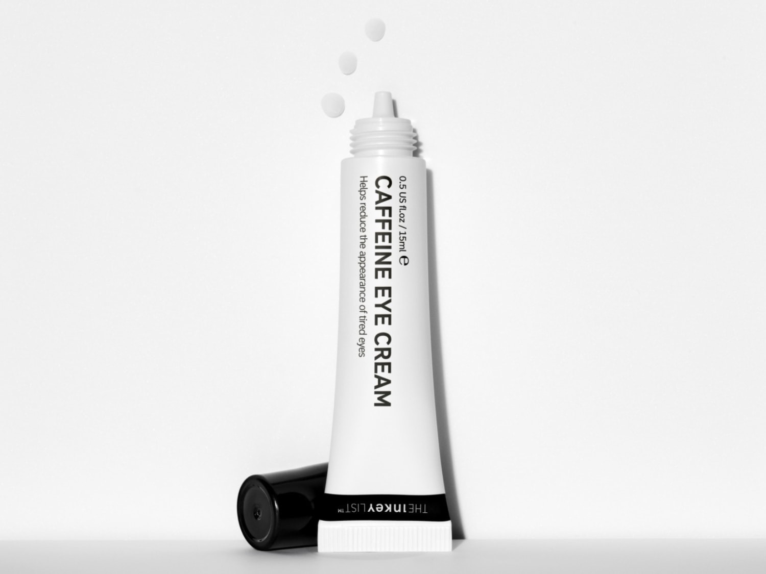 De Inkey List Caffeine Eye Cream in een witte tube met zwarte dop, met drie druppels crème die de vermindering van vermoeide ogen beloven.