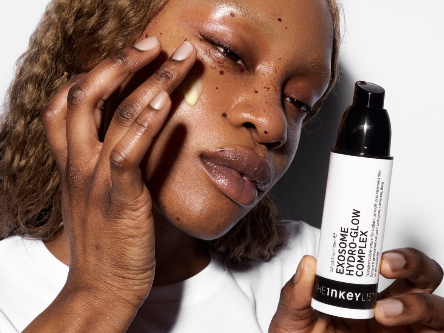Een vrouw met donkere huidskleur brengt een lichtgele serum aan op haar gezicht, terwijl ze een witte fles The Inkey List Exosome Hydro-Glow Complex vasthoudt.