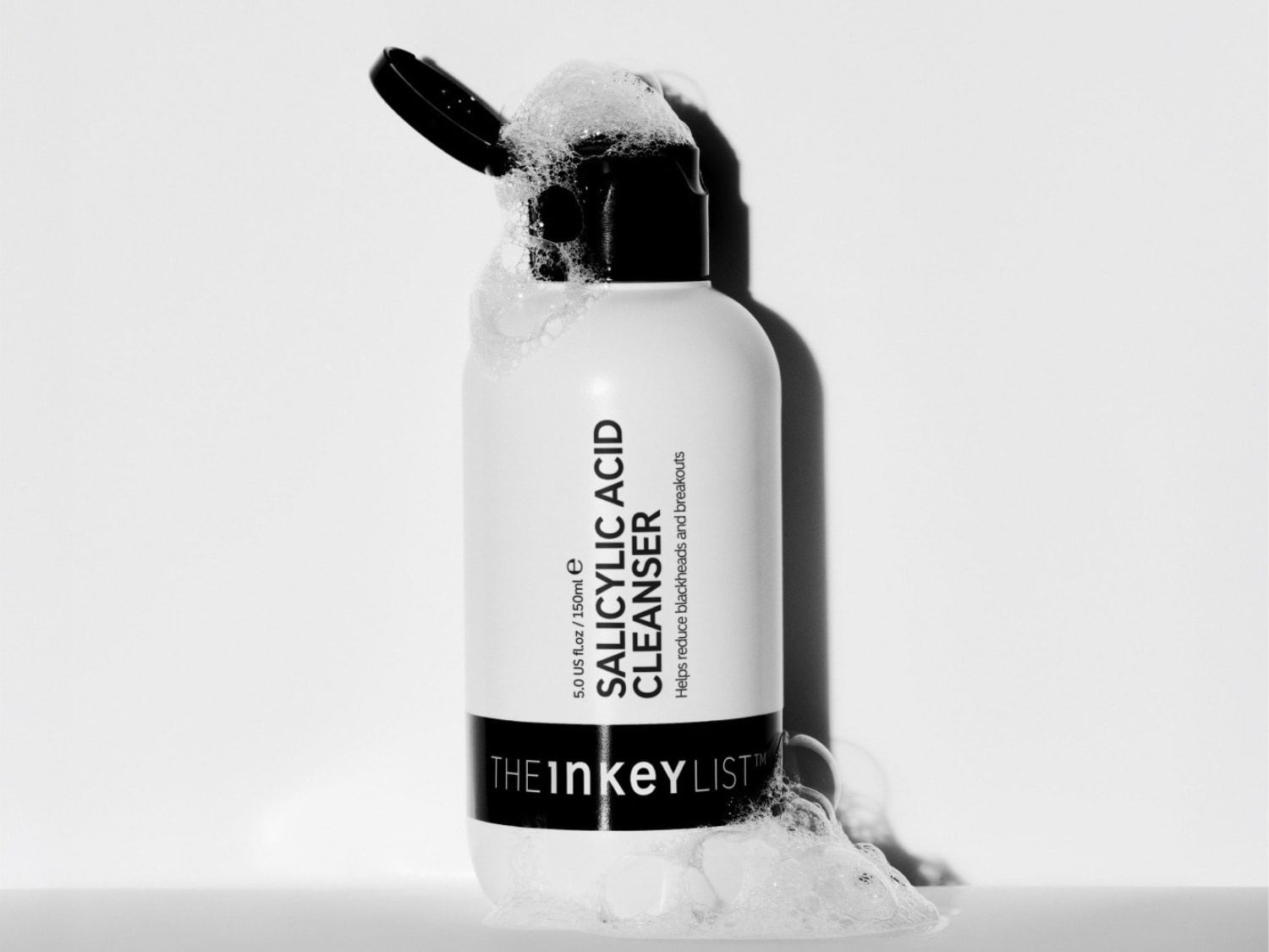 De witte fles van The Inkey List Salicylic Acid Cleanser, met een zwarte dop en schuim dat over de fles en de ondergrond stroomt, helpt mee-eters en puistjes te verminderen.