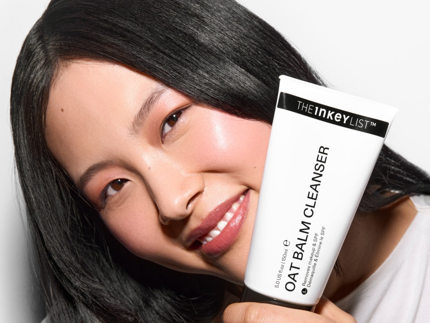 Een lachende vrouw met glanzend zwart haar houdt een witte tube THE INKEY LIST Oat Balm Cleanser vast, die make-up en SPF verwijdert.