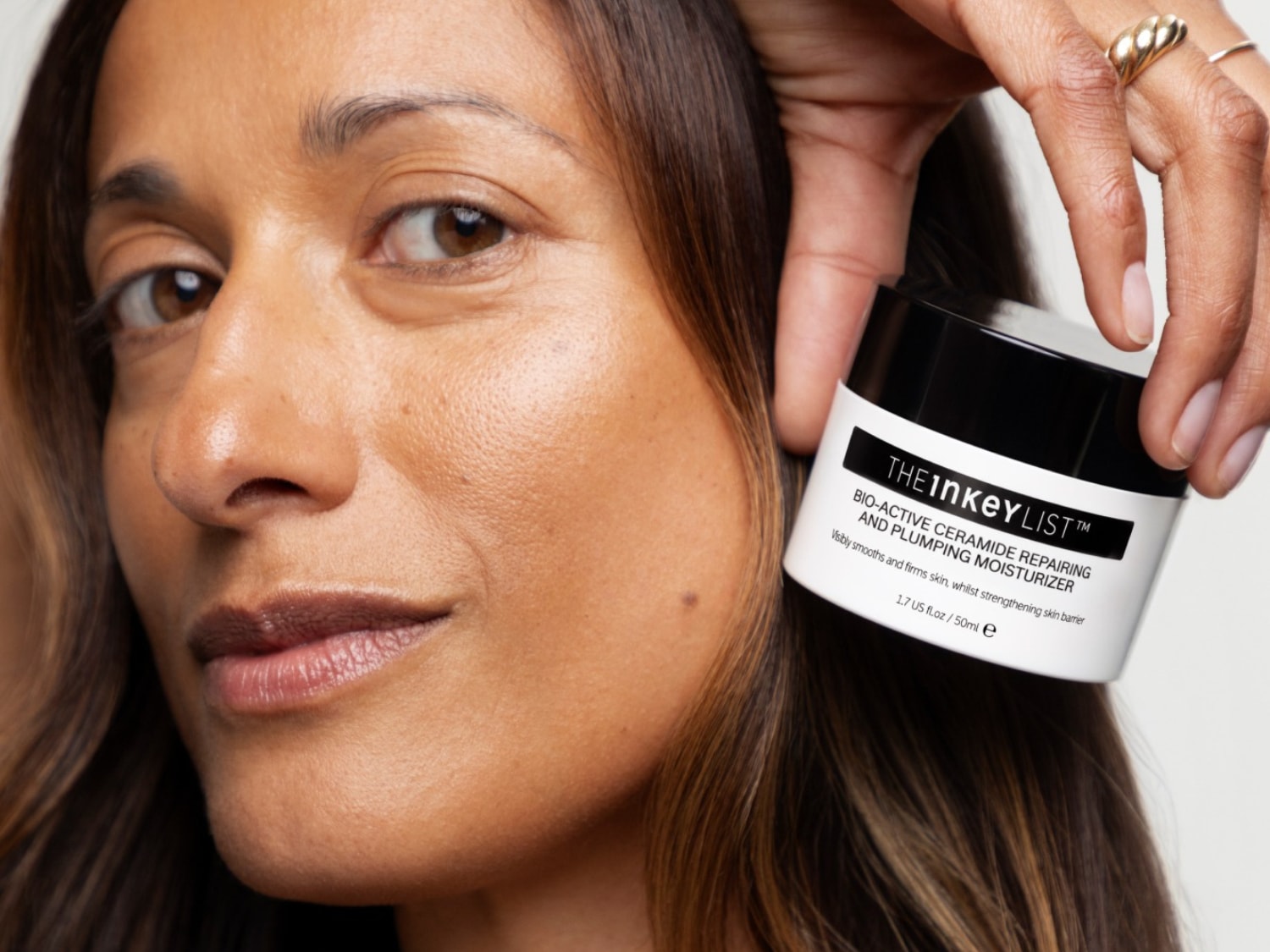 Een vrouw met bruin haar en een natuurlijke make-up look houdt een witte pot The Inkey List Bio-Active Ceramide Repairing and Plumping Moisturizer van 50 ml vast, die de huid zichtbaar gladder en steviger maakt en de huidbarrière versterkt.
