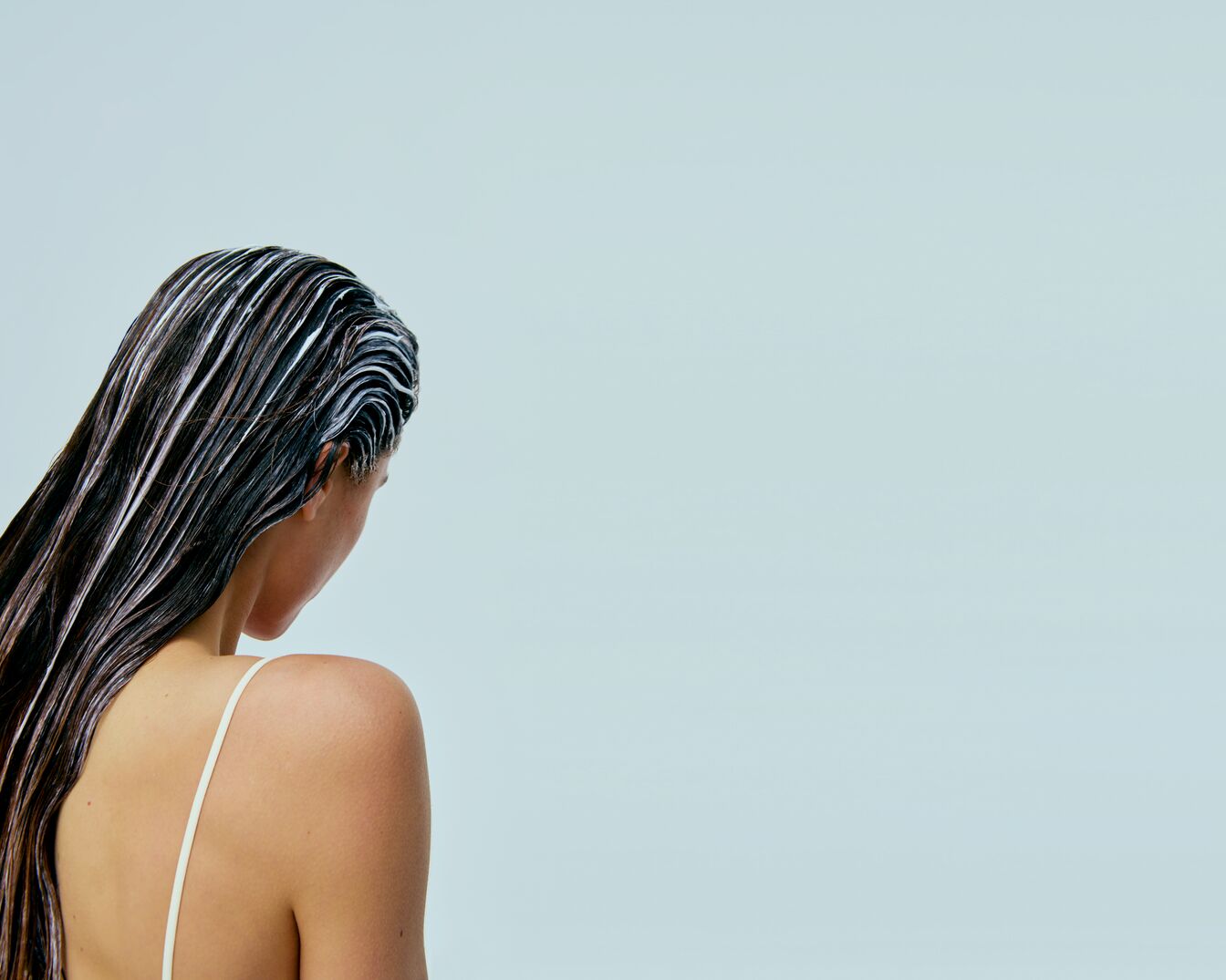 Eine Frau von hinten mit langen, dunklen Haaren, auf die eine weiße Creme aufgetragen wurde, vor einem hellblauen Hintergrund.