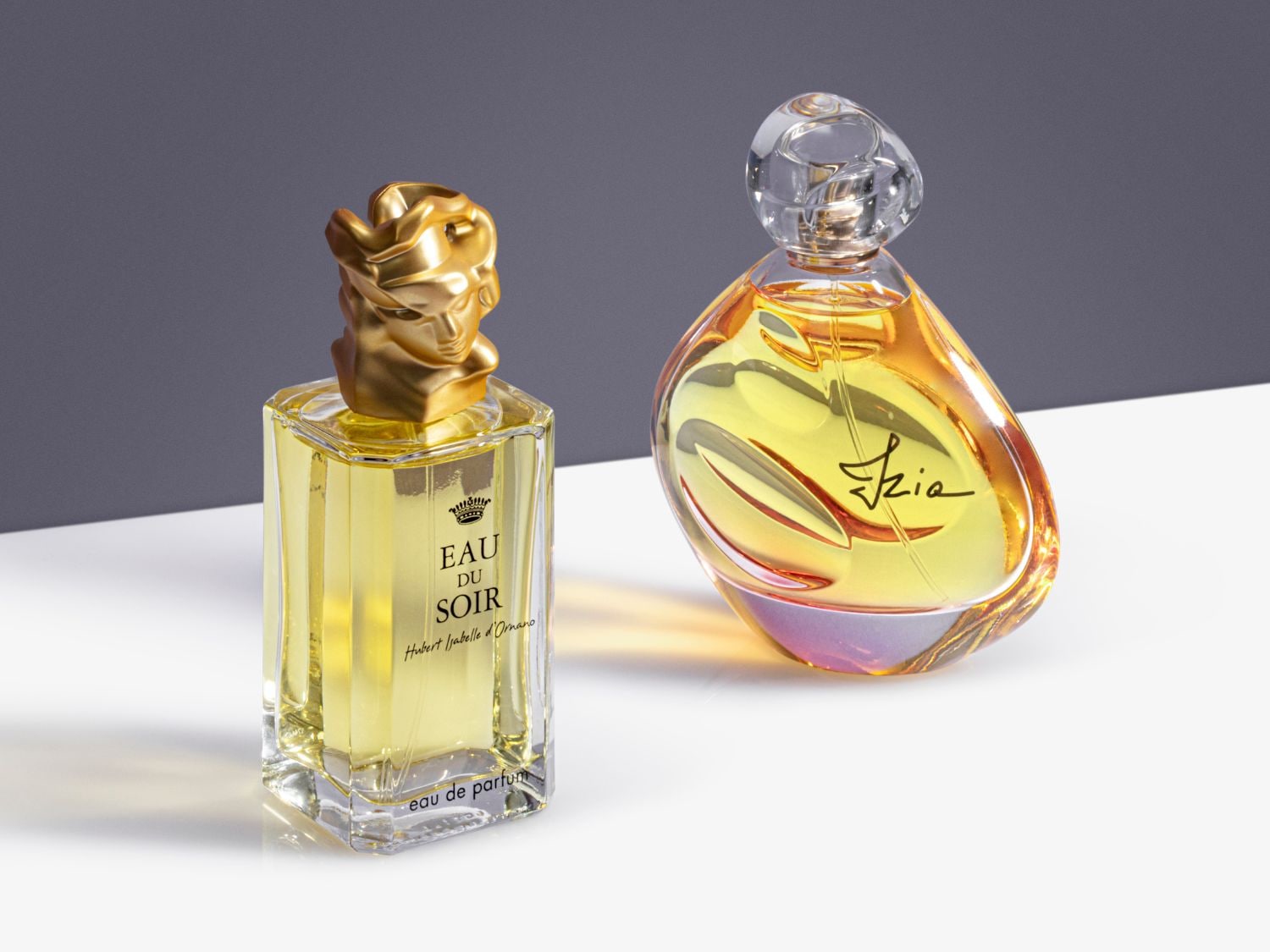 Twee flessen Eau de Parfum: 'Eau du Soir' heeft een gouden dop in de vorm van een gezicht en een heldere, rechthoekige fles, terwijl 'Izia' een afgeronde fles heeft met een heldere dop en gouden vloeistof.
