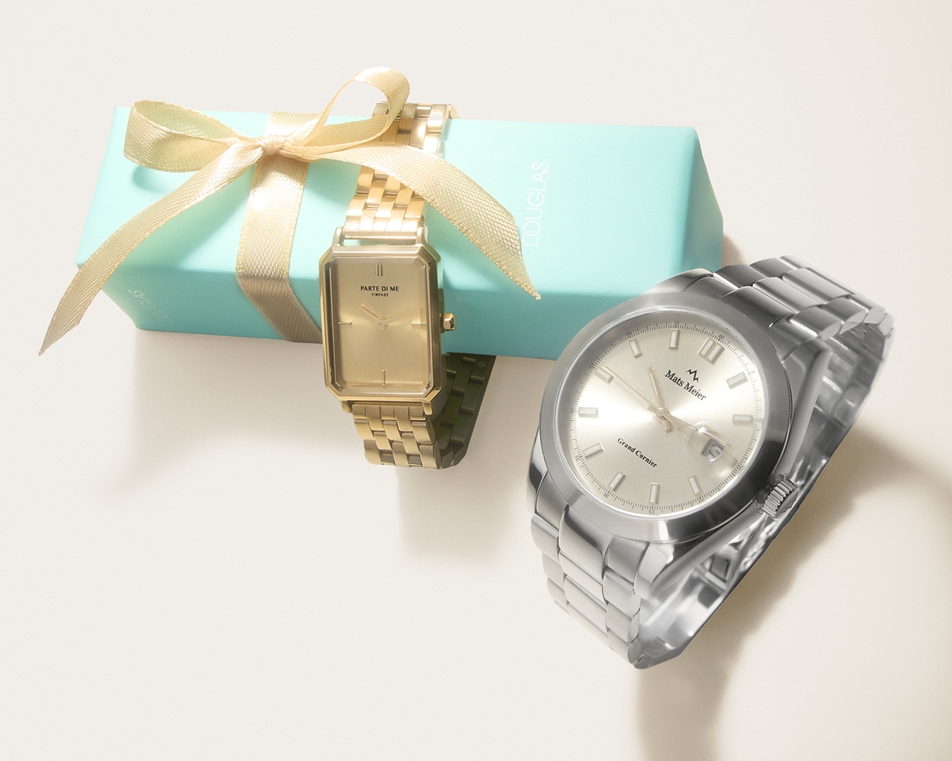 Nahaufnahme von zwei eleganten Uhren, eine goldene 'Parte di Me Firenze' und eine silberne 'Mats Meier Grand Corner', neben einer hellblauen Douglas-Geschenkbox mit goldener Schleife.