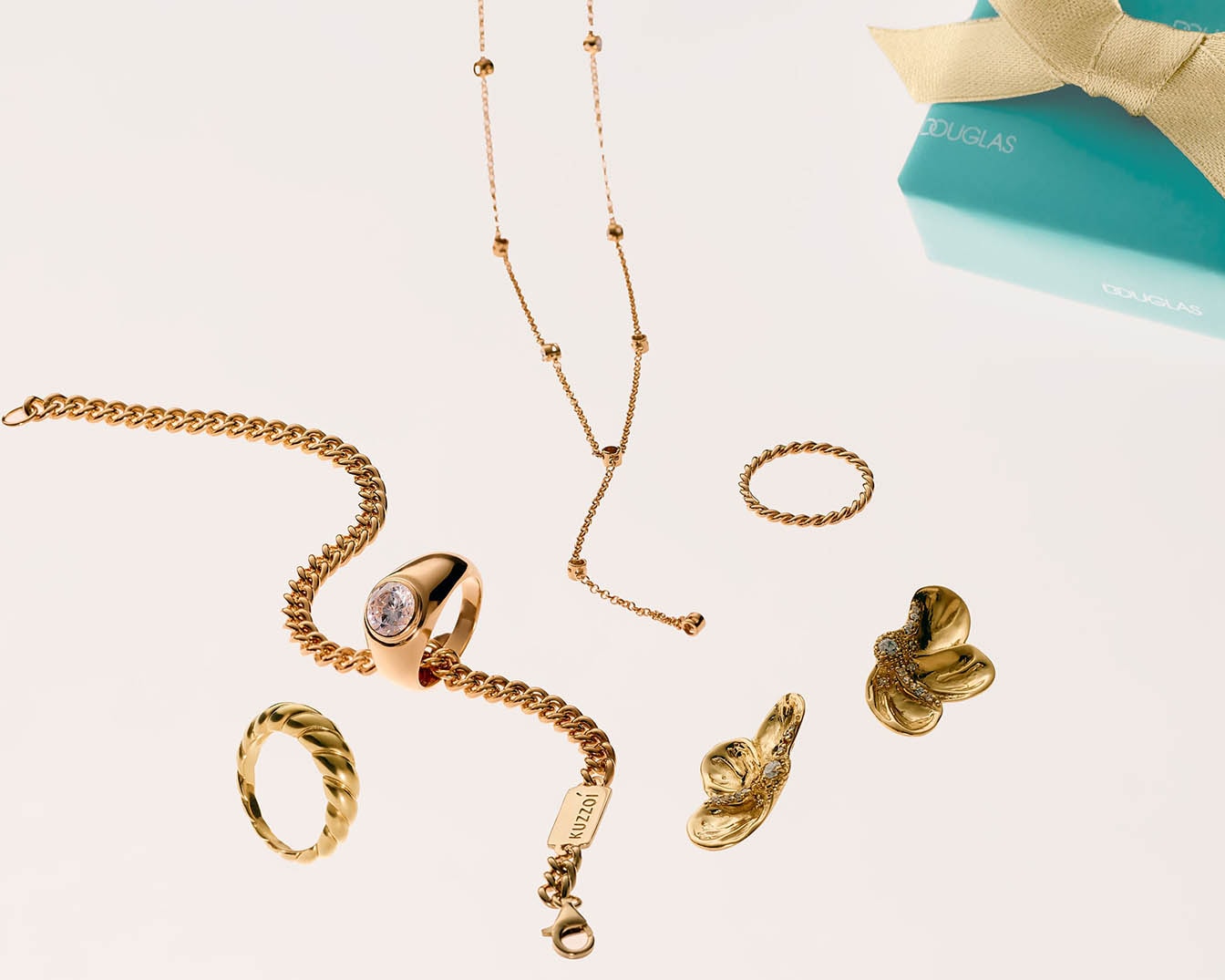 Entdecken Sie goldfarbene Accessoires wie Ringe, Halsketten und Armbänder, die auf einem hellen Hintergrund mit einer türkisfarbenen Douglas-Box präsentiert werden.