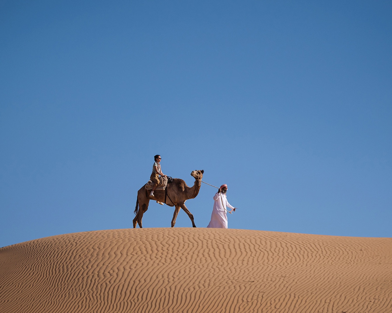 Sotto un cielo azzurro, una persona a cavallo di un cammello marrone è guidata da un uomo in abito tradizionale bianco su una duna di sabbia dorata.