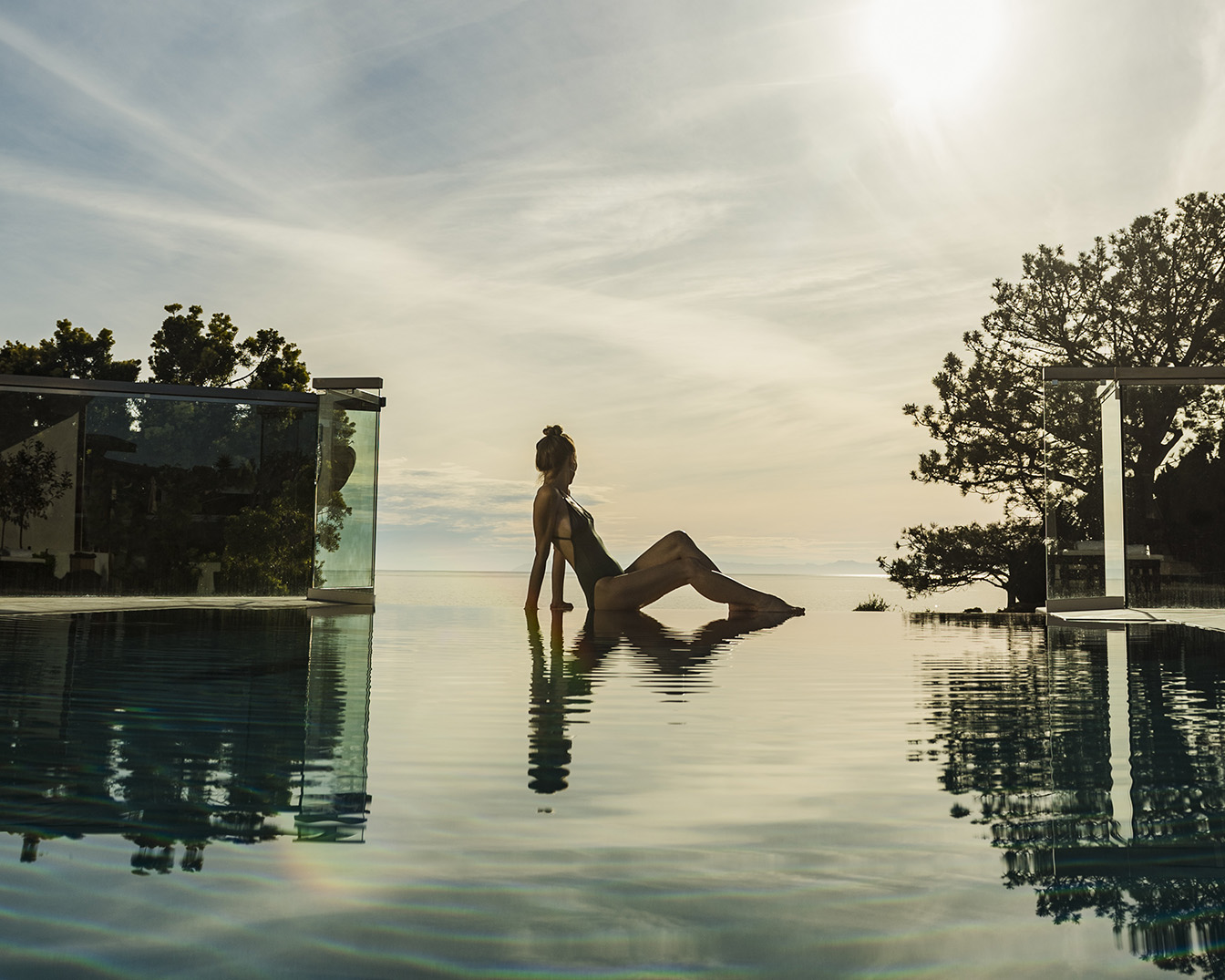 Una donna in costume da bagno è seduta sul bordo di una piscina a sfioro, con lo sguardo rivolto verso l'orizzonte e il cielo sereno.