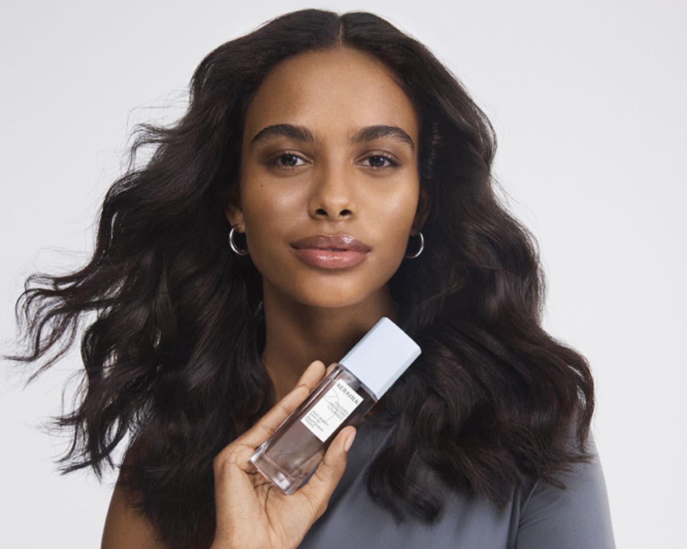 Een vrouw met golvend donker haar houdt een heldere Kerastase-fles vast met een lichtblauwe dop, wat de focus op haarverzorging benadrukt.
