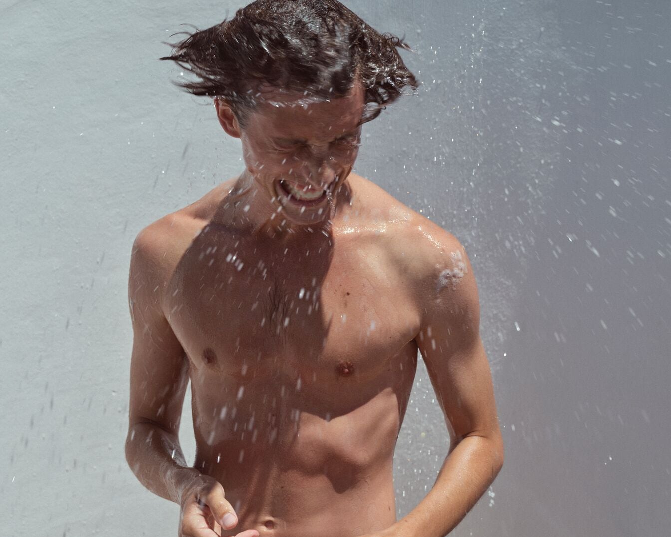 Ein Mann mit nacktem Oberkörper steht im Wasser und lacht, während er von Wassertropfen umgeben ist.
