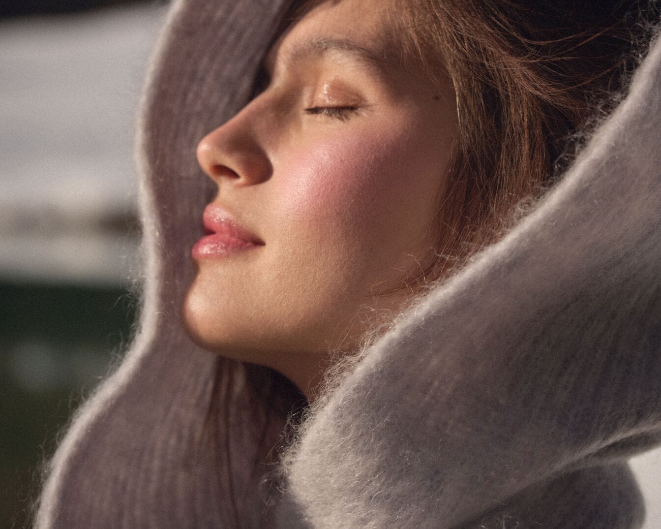 Nahaufnahme einer Frau mit geschlossenen Augen, deren Gesicht von einem weichen, hellgrauen Stoff umrahmt wird, der ihre rosigen Wangen und glänzenden Lippen betont.