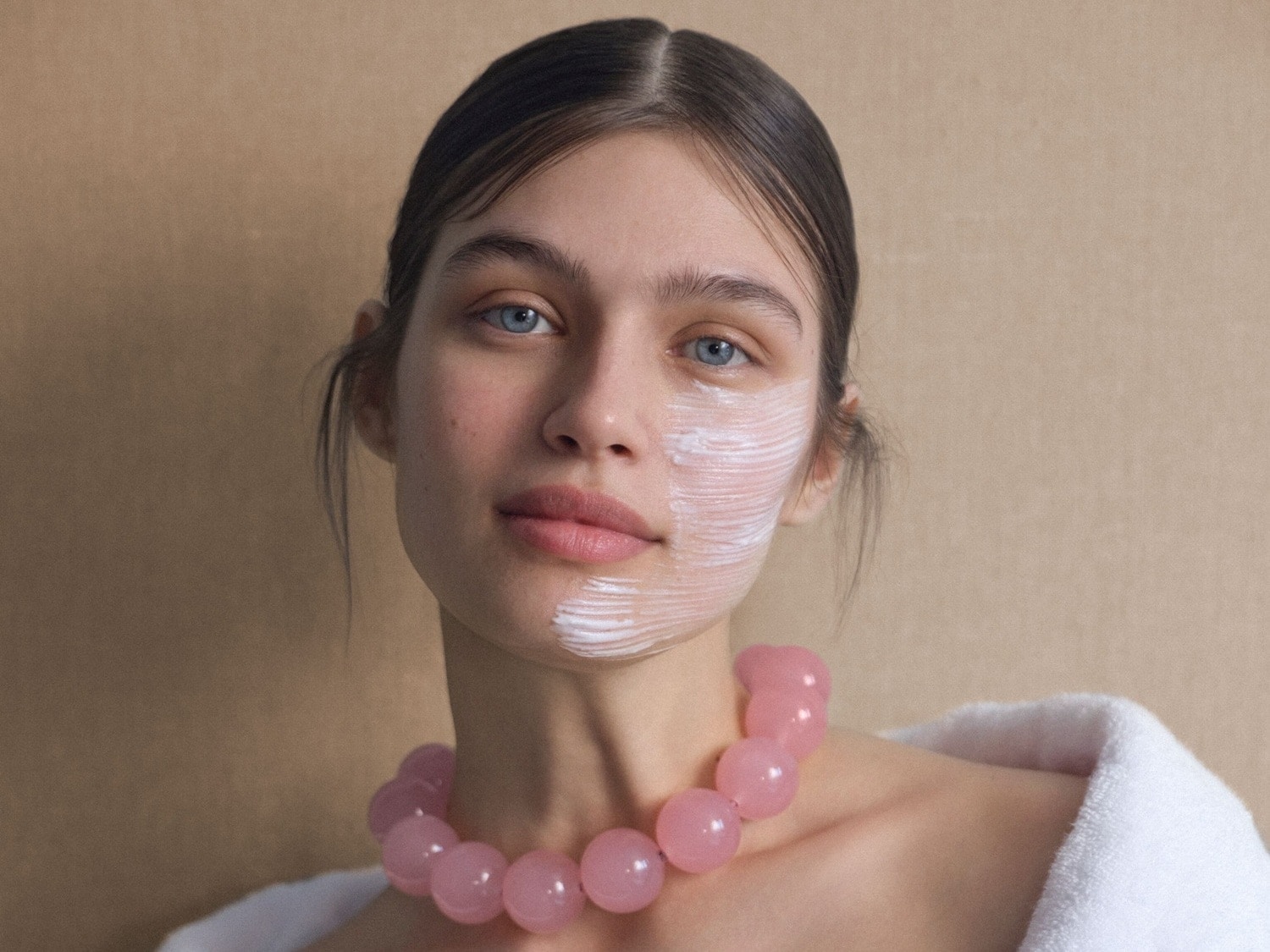 Eine Frau mit hellblauen Augen und einem weißen Bademantel trägt eine rosa Kugelkette und hat eine weiße Creme auf einer Gesichtshälfte.