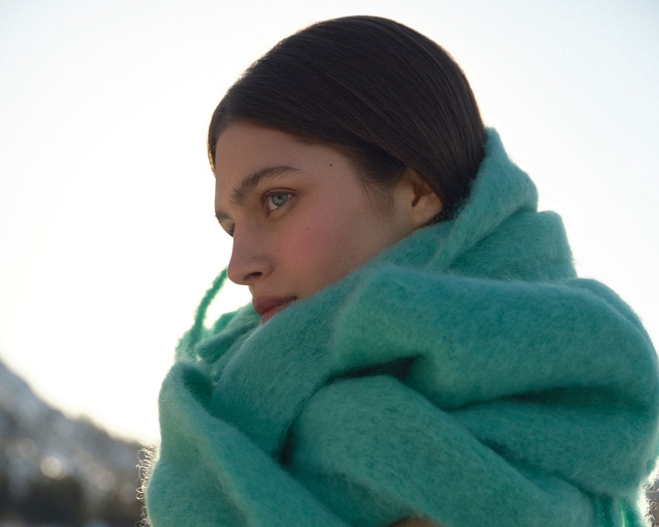 Nahaufnahme einer Frau mit hellblauen Augen und braunem Haar, die in einen flauschigen, helltürkisfarbenen Schal gehüllt ist.