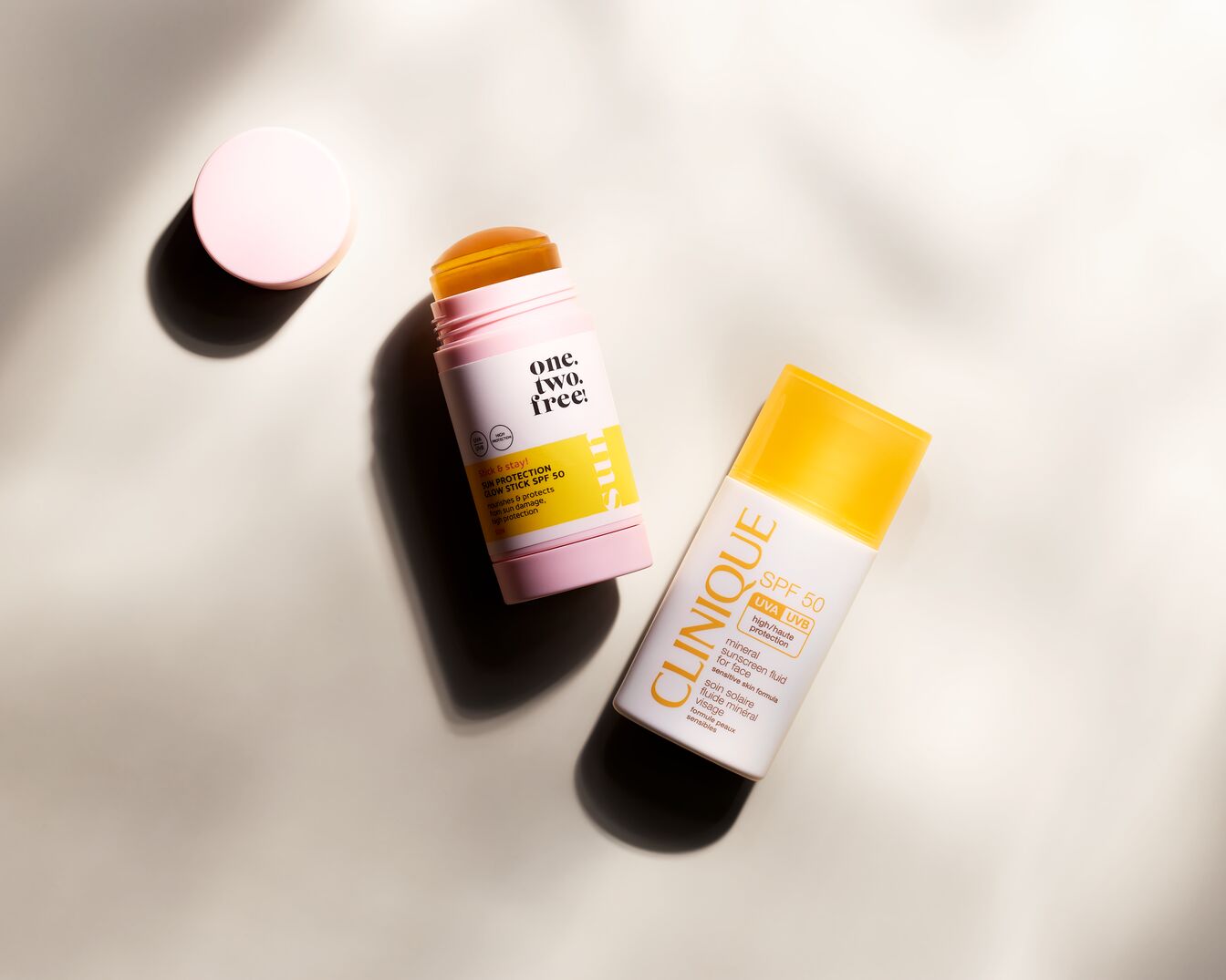 Sonnenpflege-Duo: One.Two.Free! Glow Stick SPF 50 in rosa Verpackung und Clinique Mineral Sunscreen Fluid SPF 50 in weißer Flasche mit gelbem Deckel.