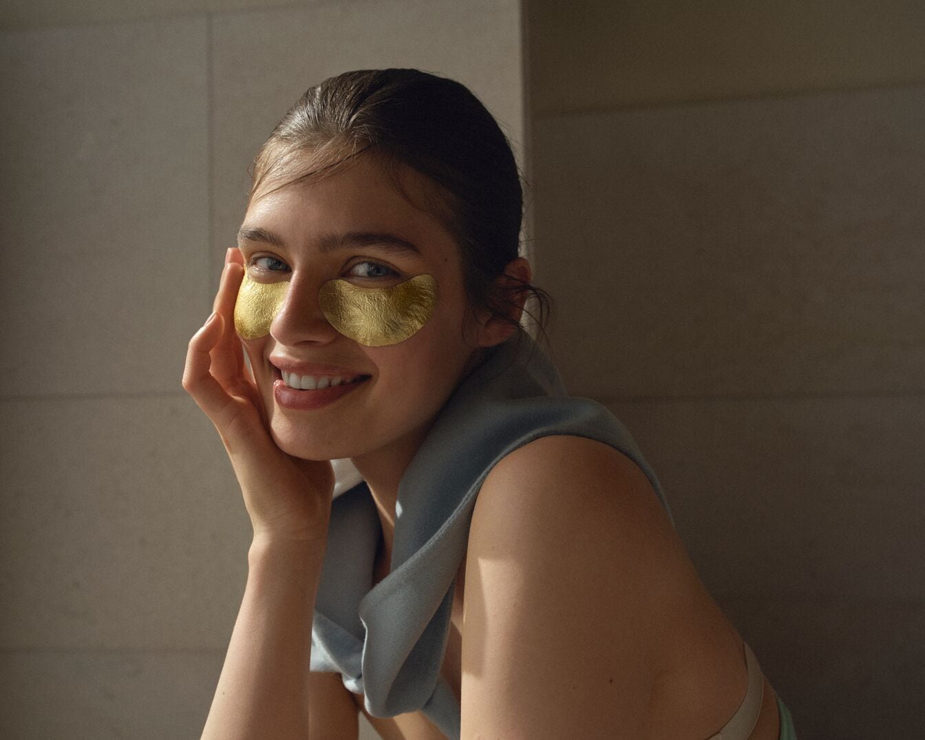 Eine lächelnde Frau mit goldenen Augenpads unter den Augen, die ihre Haut pflegen und ihr einen strahlenden Teint verleihen.