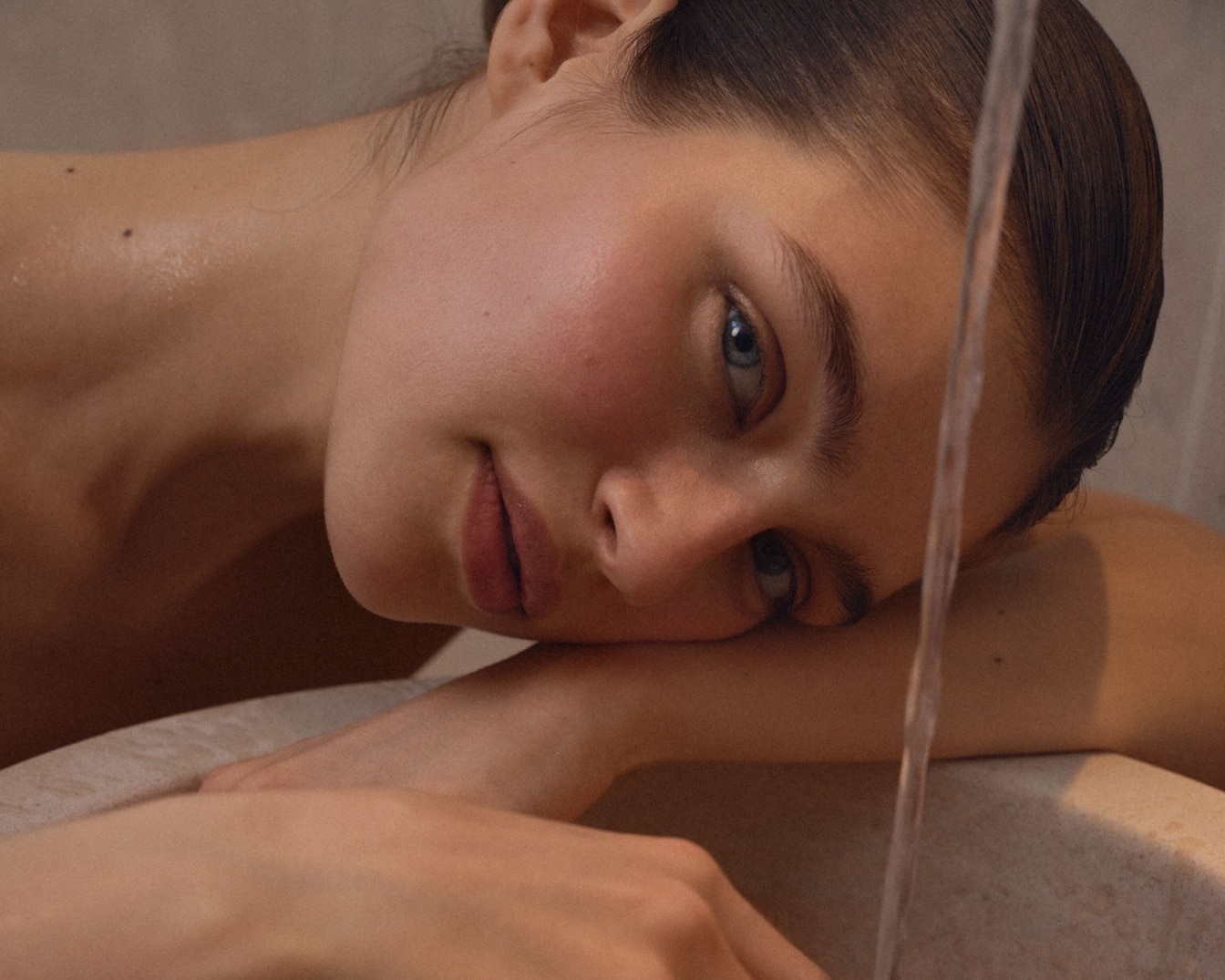 Nahaufnahme einer Frau mit hellblauen Augen, die ihr Gesicht auf ihre Hände stützt, während ein Wasserstrahl über ihr Gesicht fließt.