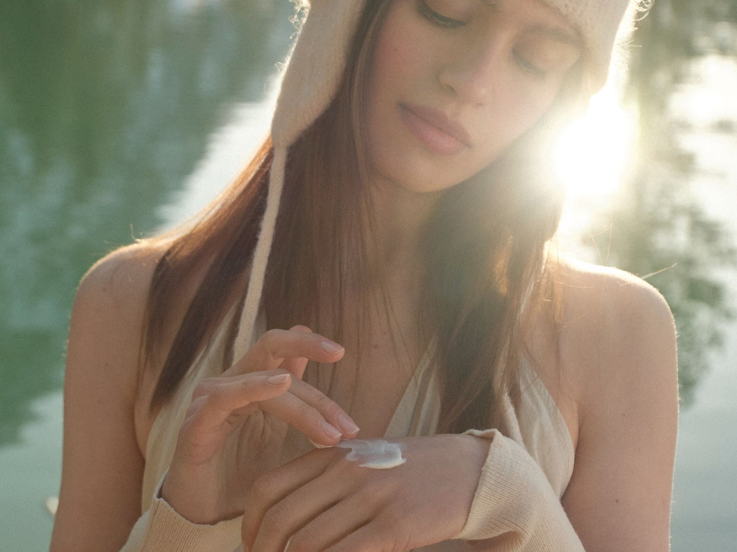 Eine Frau mit hellbraunem Haar und einem cremefarbenen Hut trägt eine Feuchtigkeitscreme auf ihre Hand auf, während sie in der Sonne sitzt.