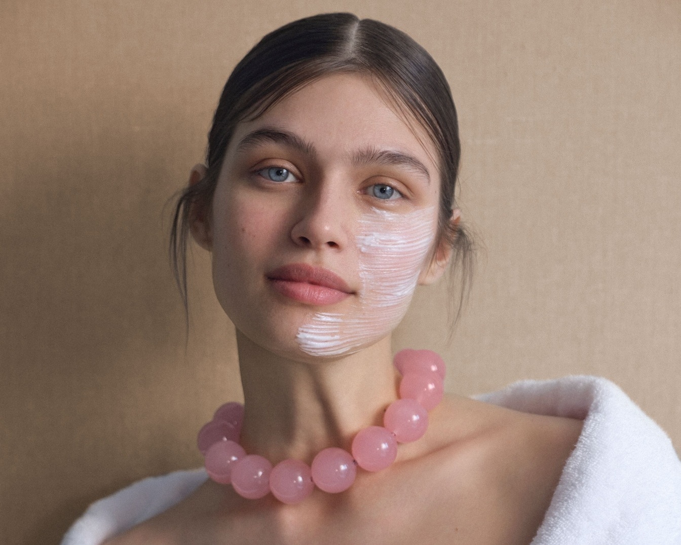 Eine Frau mit blauen Augen und weißer Gesichtsmaske, die eine rosa Kugelkette und einen weißen Bademantel trägt.